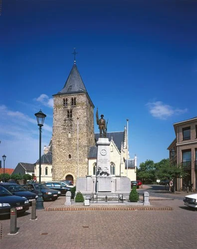 De kerk van Wellen met een toren en een standbeeld in een plein, omringd door auto's en gebouwen, onder een blauwe hemel.