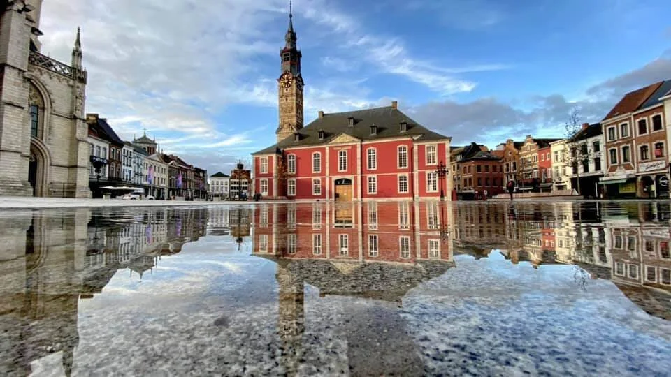 Het marktplein van Sint-Truiden gefotografeerd met een reflectie van de regen. In het midden het stadhuis met belforttoren, omringd door kleurrijke gebouwen onder een blauwe lucht.