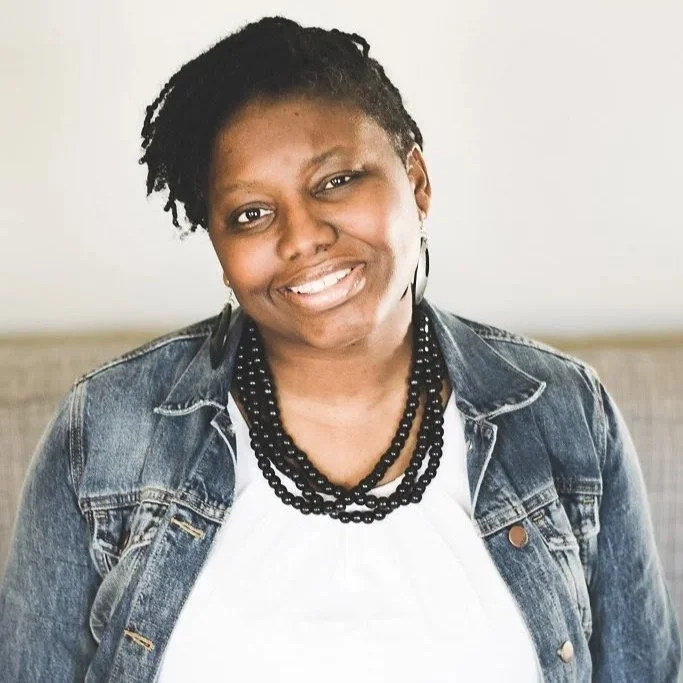 A woman with short curly hair, smiling, wearing a denim jacket, black beaded necklaces, and a white top, sitting indoors against a plain background.