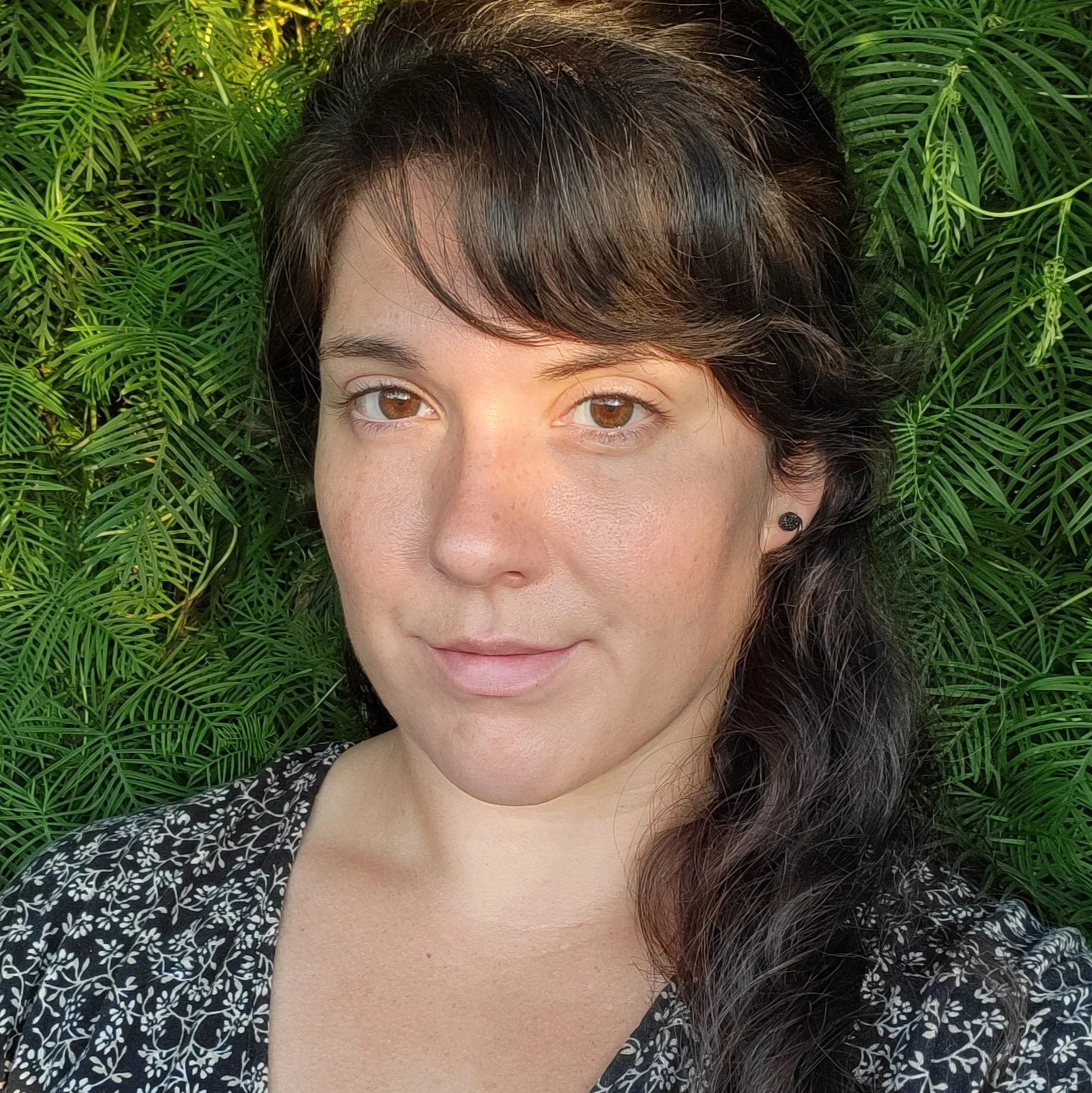 A woman with dark hair and brown eyes lying on green fern-like foliage, wearing a black and white patterned top and small earrings.