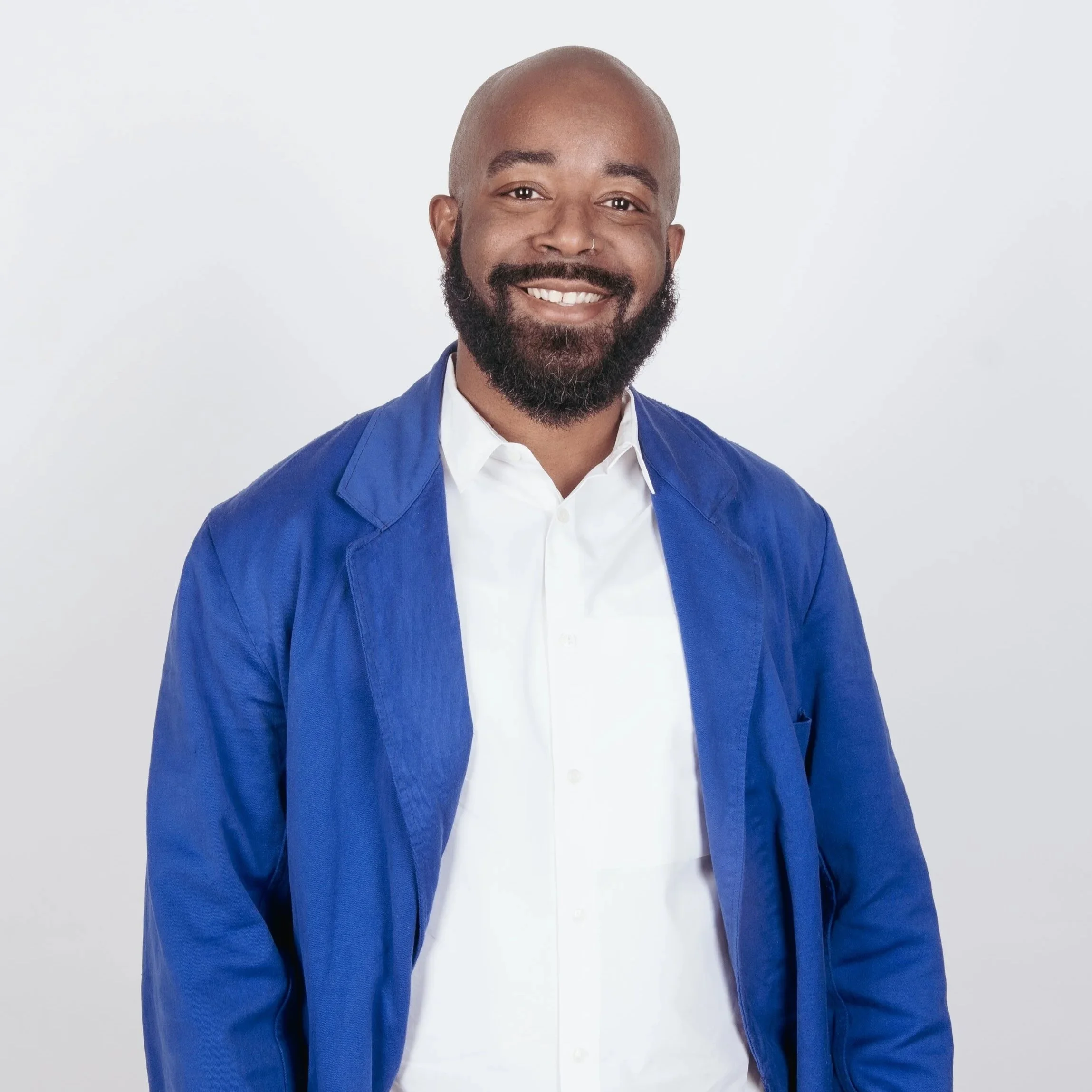 A smiling Black man with a beard, mustache, and bald head, wearing a white shirt and a blue blazer, standing against a plain white background.