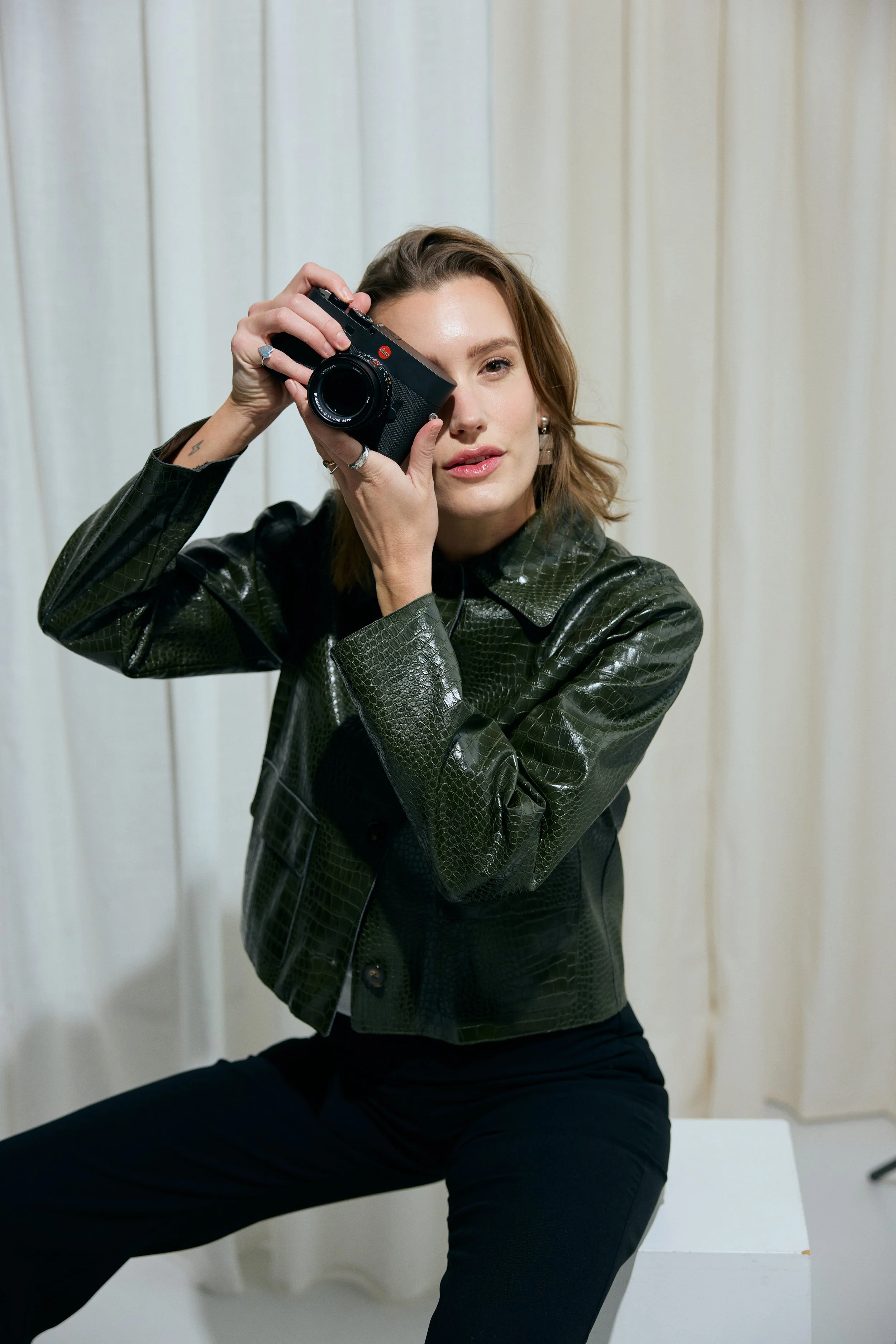 A woman with shoulder-length brown hair holding a black camera up to her eye, sitting on a white surface in front of a white curtain, wearing a black leather jacket with a snake pattern, a black top, and black pants.
