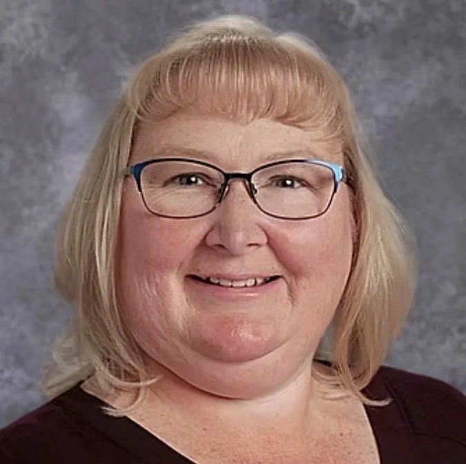A woman with blonde hair, glasses, and a friendly smile, wearing a dark top, posed against a neutral background.