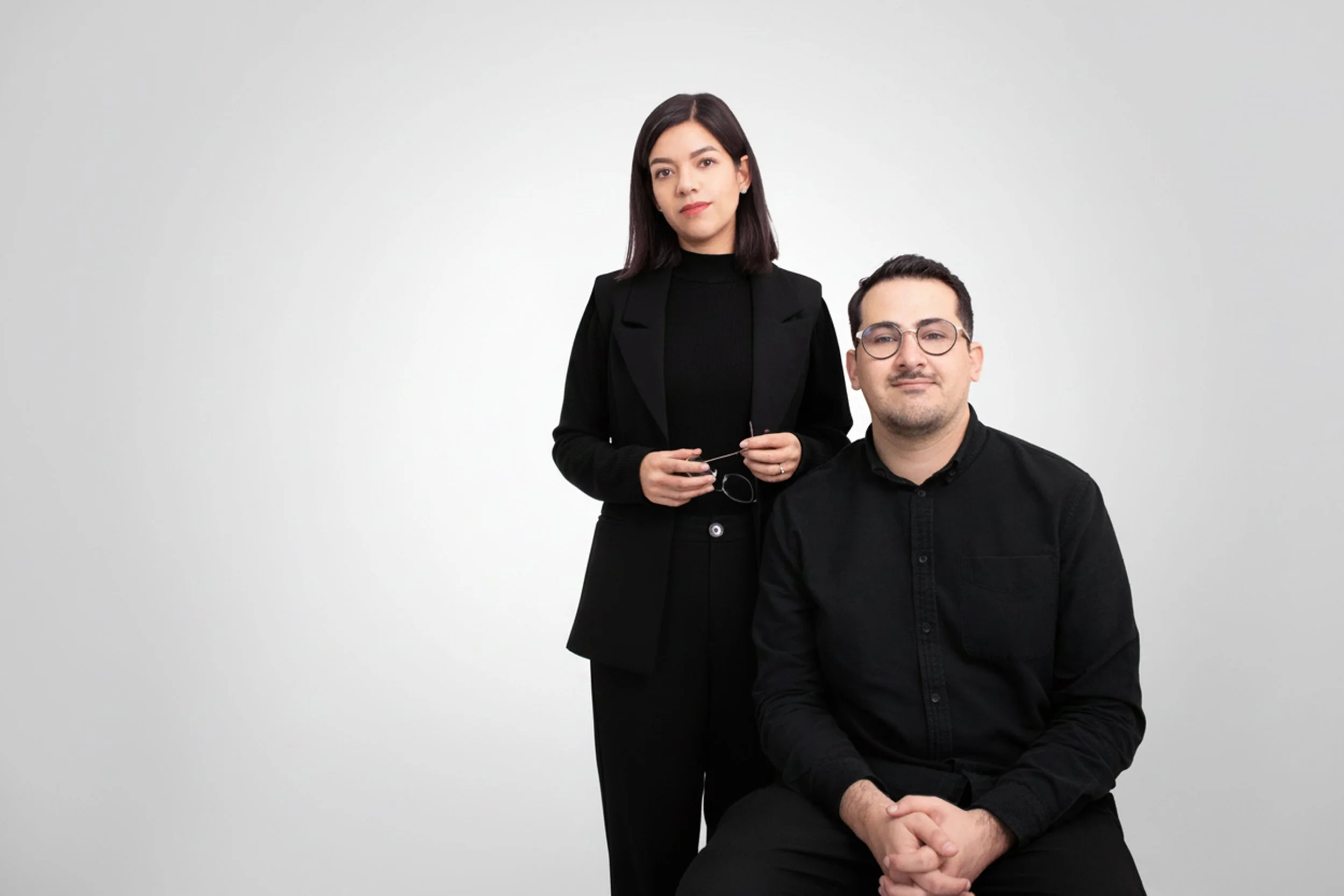 Hombre y mujer con ropa negra posando frente a fondo blanco, ella de pie sosteniendo gafas, él sentado con las manos juntas.