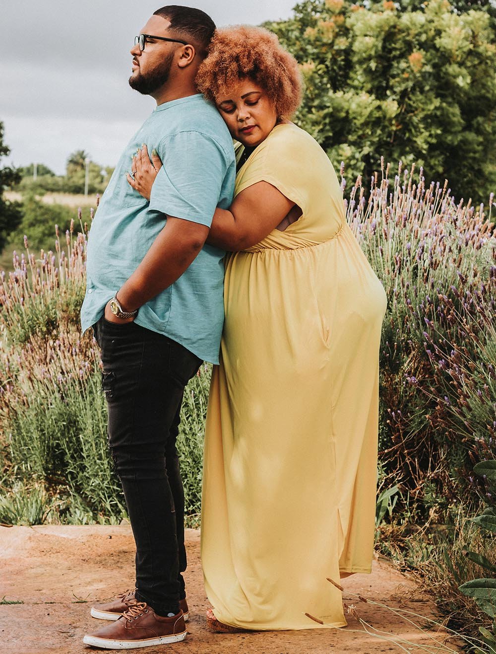 A couple standing outdoors in a field of lavender, hugging each other with eyes closed, expressing affection and serenity.