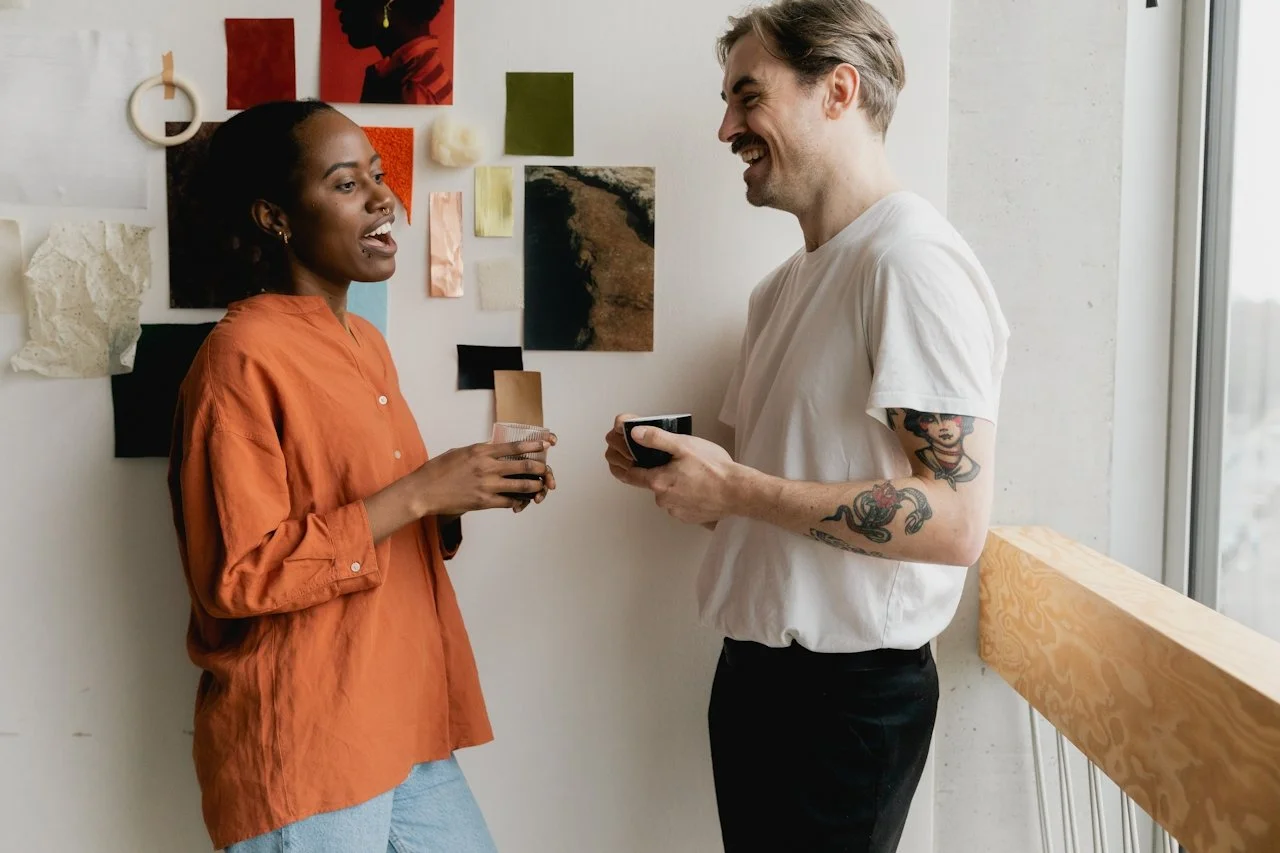 A woman in an orange blouse and a man in a white t-shirt with tattoos on his arm are smiling and holding coffee mugs while talking in a modern room with abstract art on the wall and a large window.