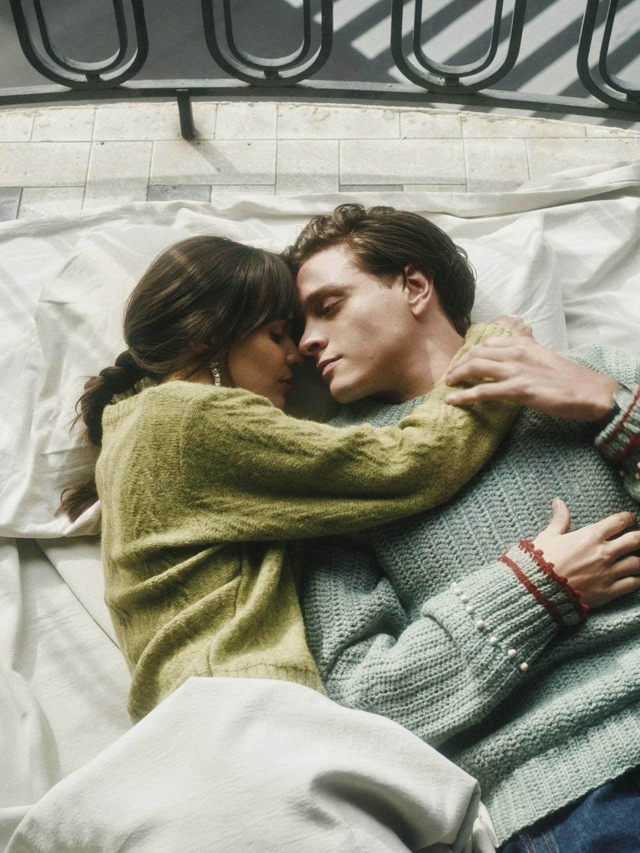A young couple is lying on a bed, embracing each other with their eyes closed, appearing peaceful and intimate. The woman has dark hair and is wearing a green sweater, while the man has brown hair and is wearing a light blue sweater.