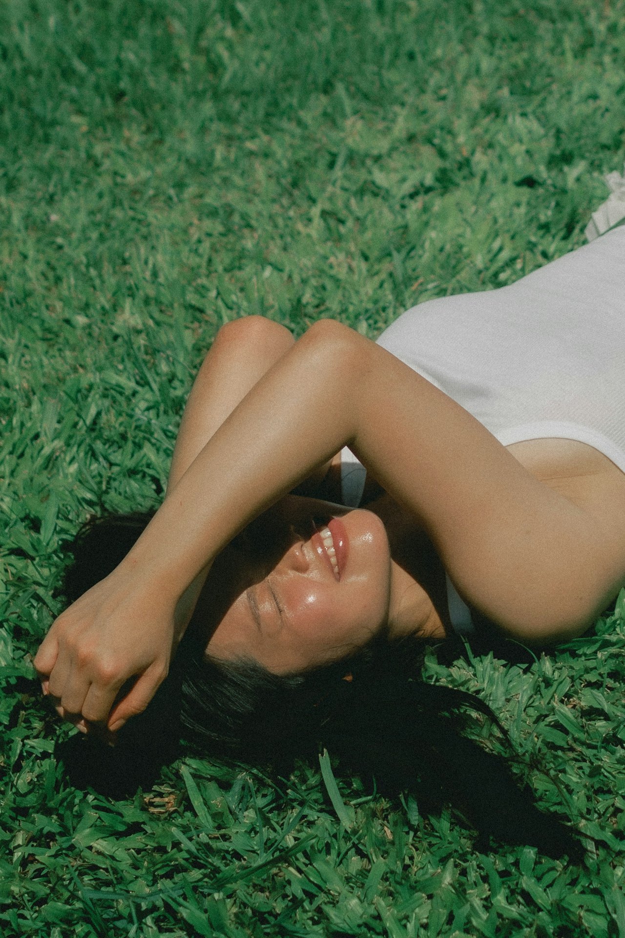 A woman lying on green grass, smiling, with one arm over her face and her head turned to the side, wearing a white top and sunglasses.