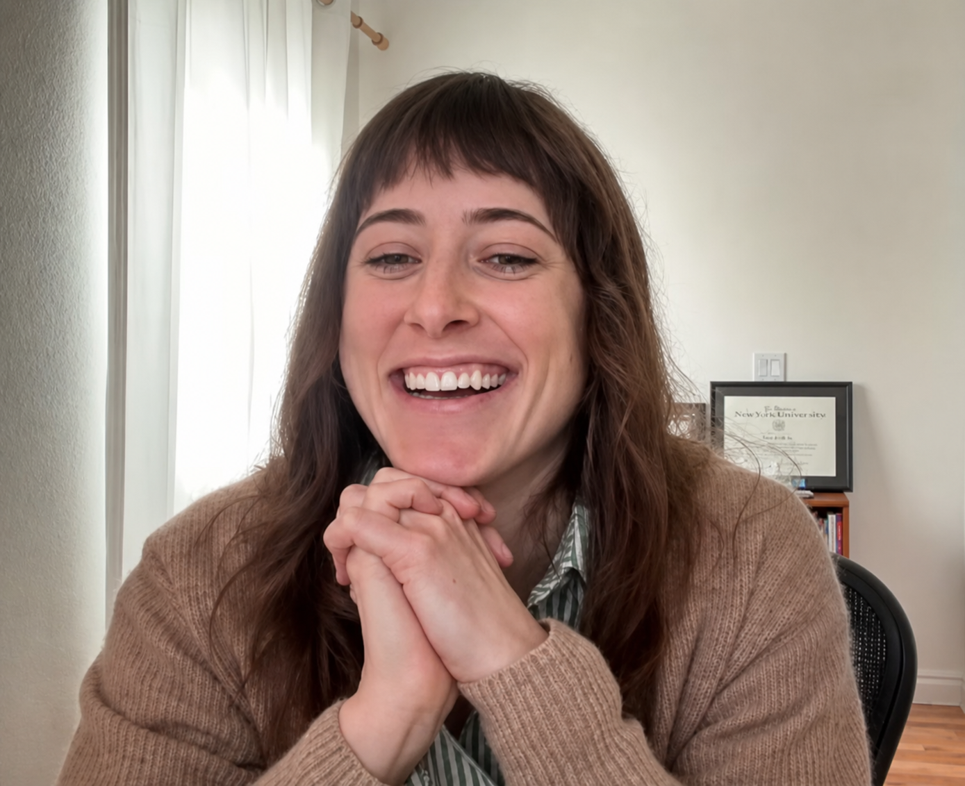 Lillian Bailey, sex therapist, Woman with brown hair and bangs smiling, resting her chin on her folded hands, in a room with a framed diploma and bookshelf in the background.