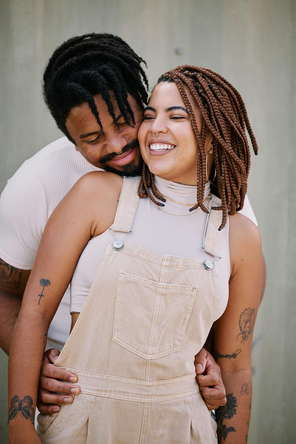 A happy couple embraces, smiling and showing affection. The woman has light brown braided hair, tattoos on her arms, and wears a beige overall with a white turtleneck. The man has black dreadlocks and a beard, leaning in close with his face near hers.
