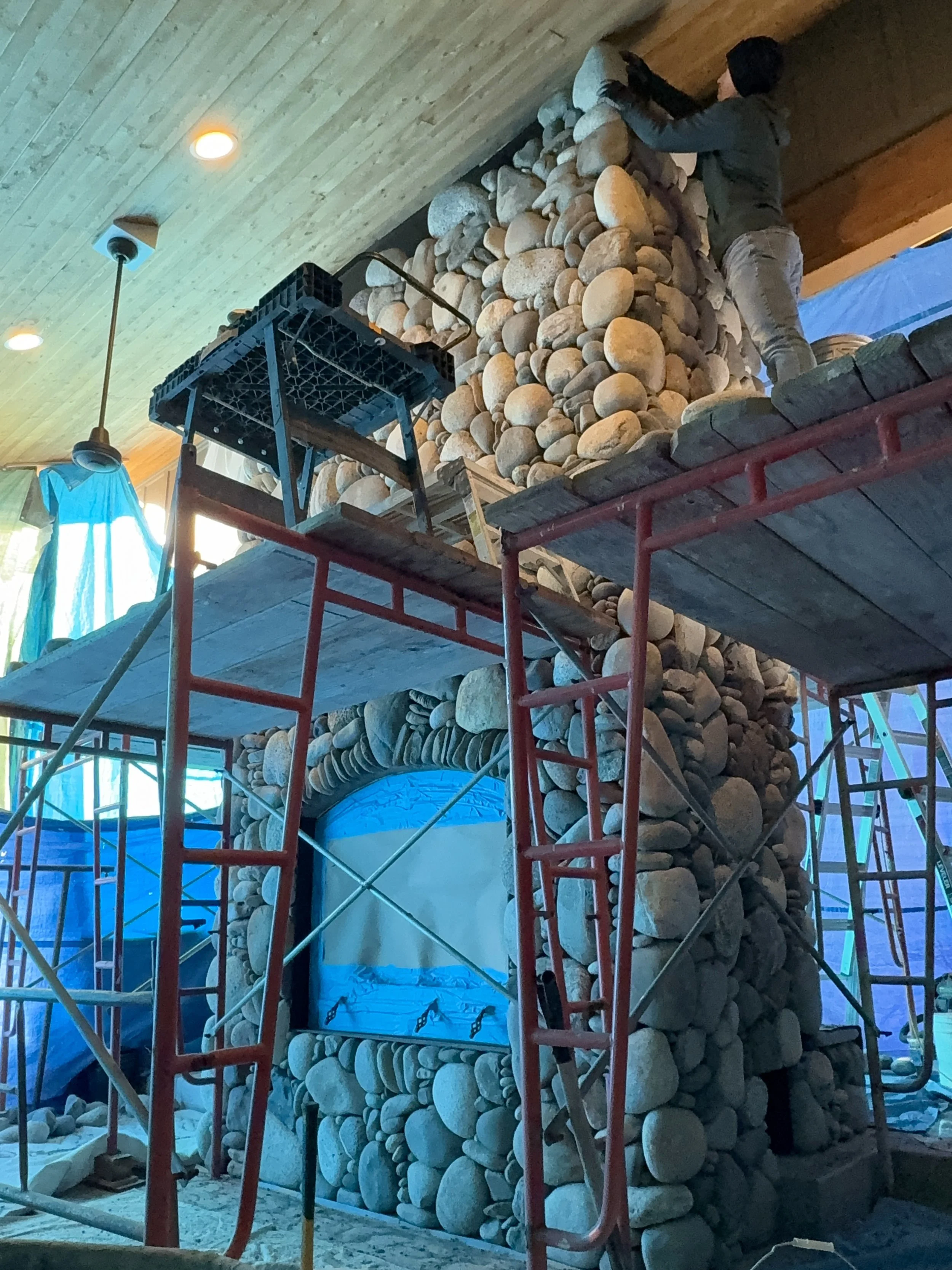 A person working on constructing a stone fireplace or wall inside a house, using a scaffold and various tools, with a wooden ceiling and windows with patterned curtains.