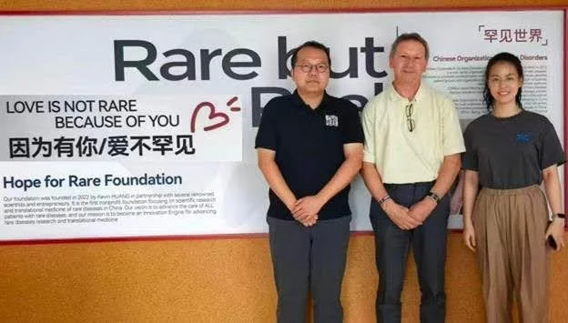 Ralf stands in between a man and a woman in front of a sign for the Hope for Rare Foundation, which has both English and Chinese text.