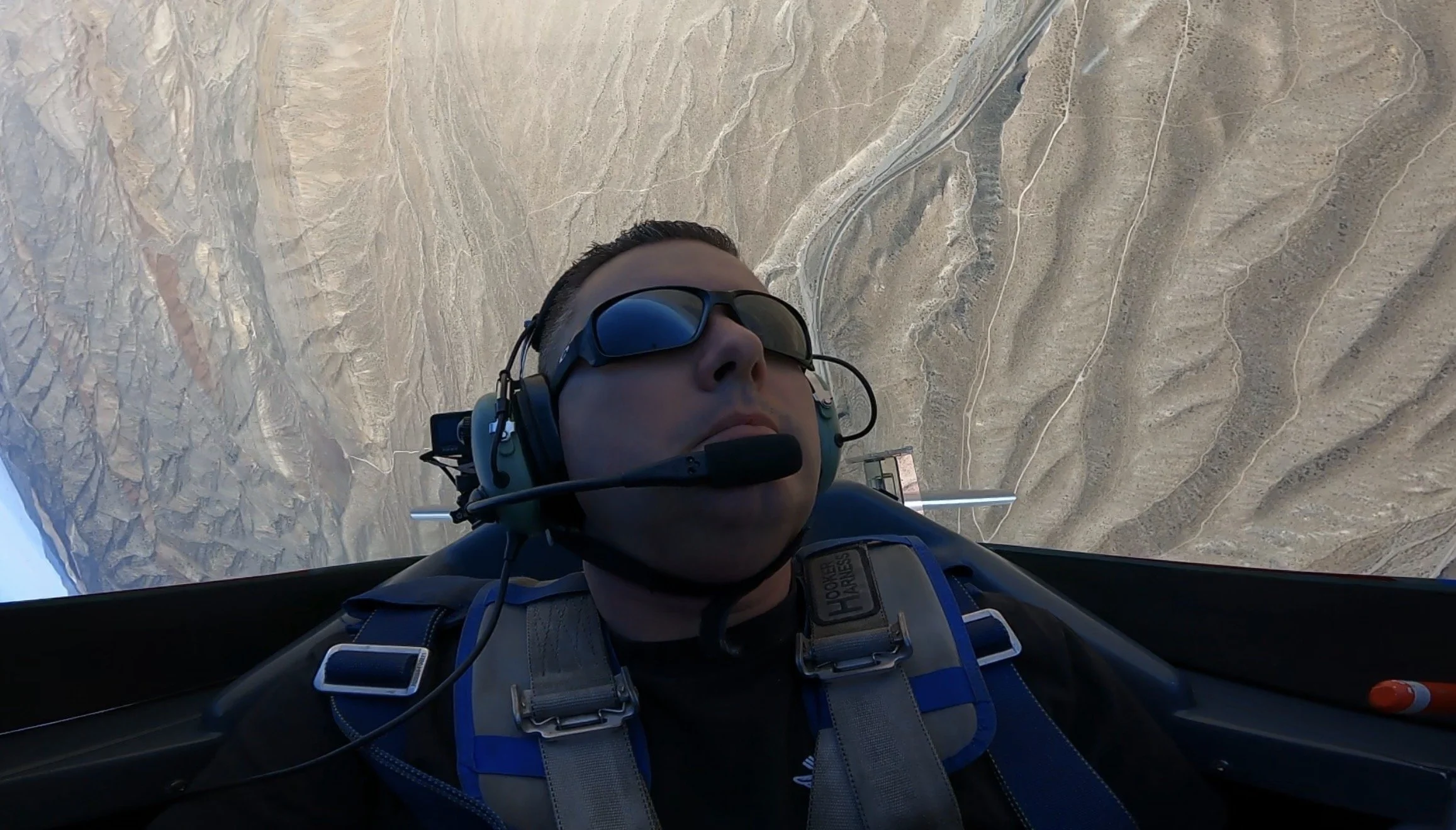 A person wearing sunglasses and a headset with microphone flying in a jet over a mountainous desert landscape.