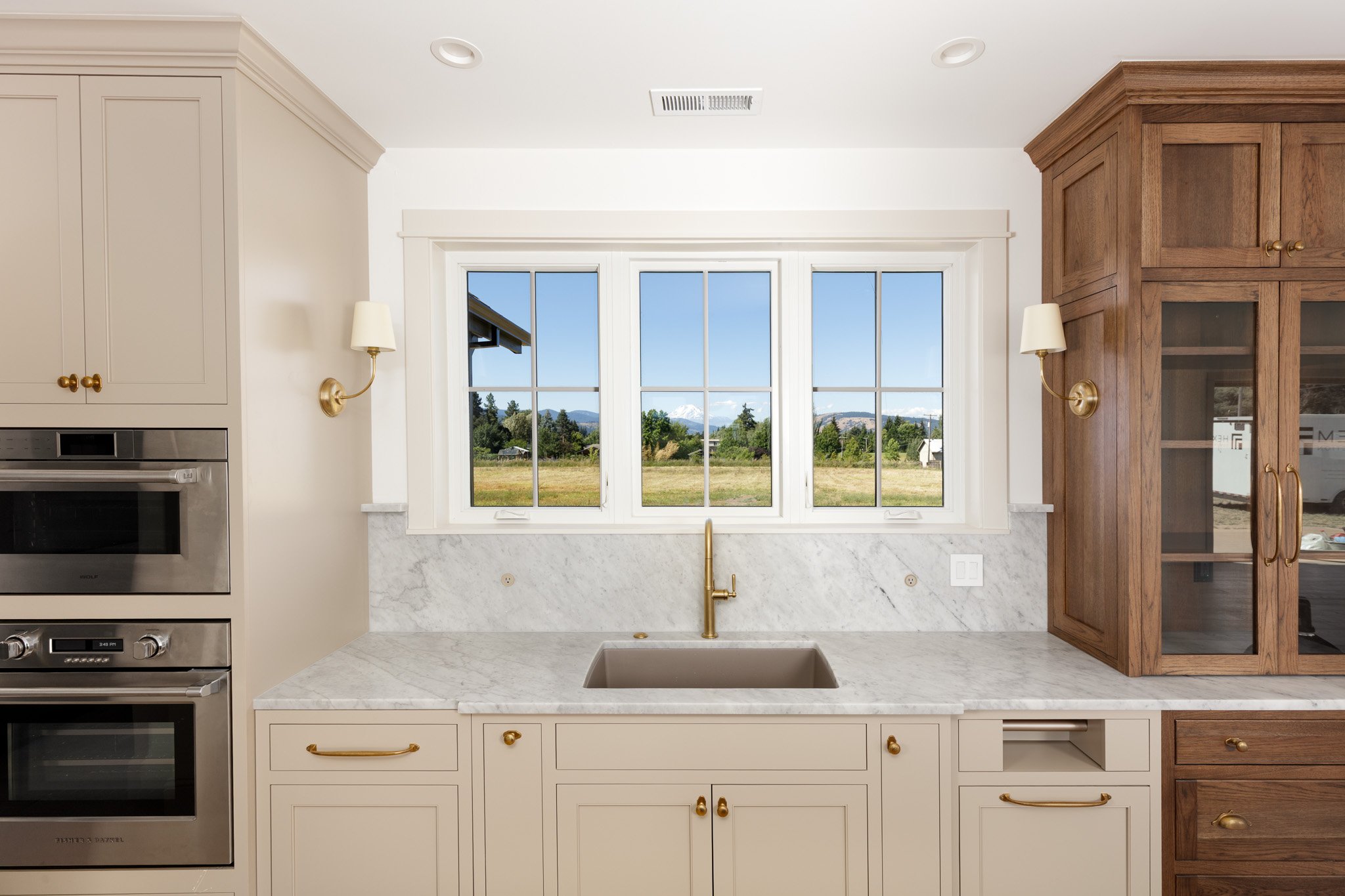 A kitchen with taupe cabinets on the left and a wooden hutch cabinet on the right. A large window shows a landscape with trees and view of Mount Adams in the distance. The countertops are carrara marble, and there is a bronze faucet over a sink.