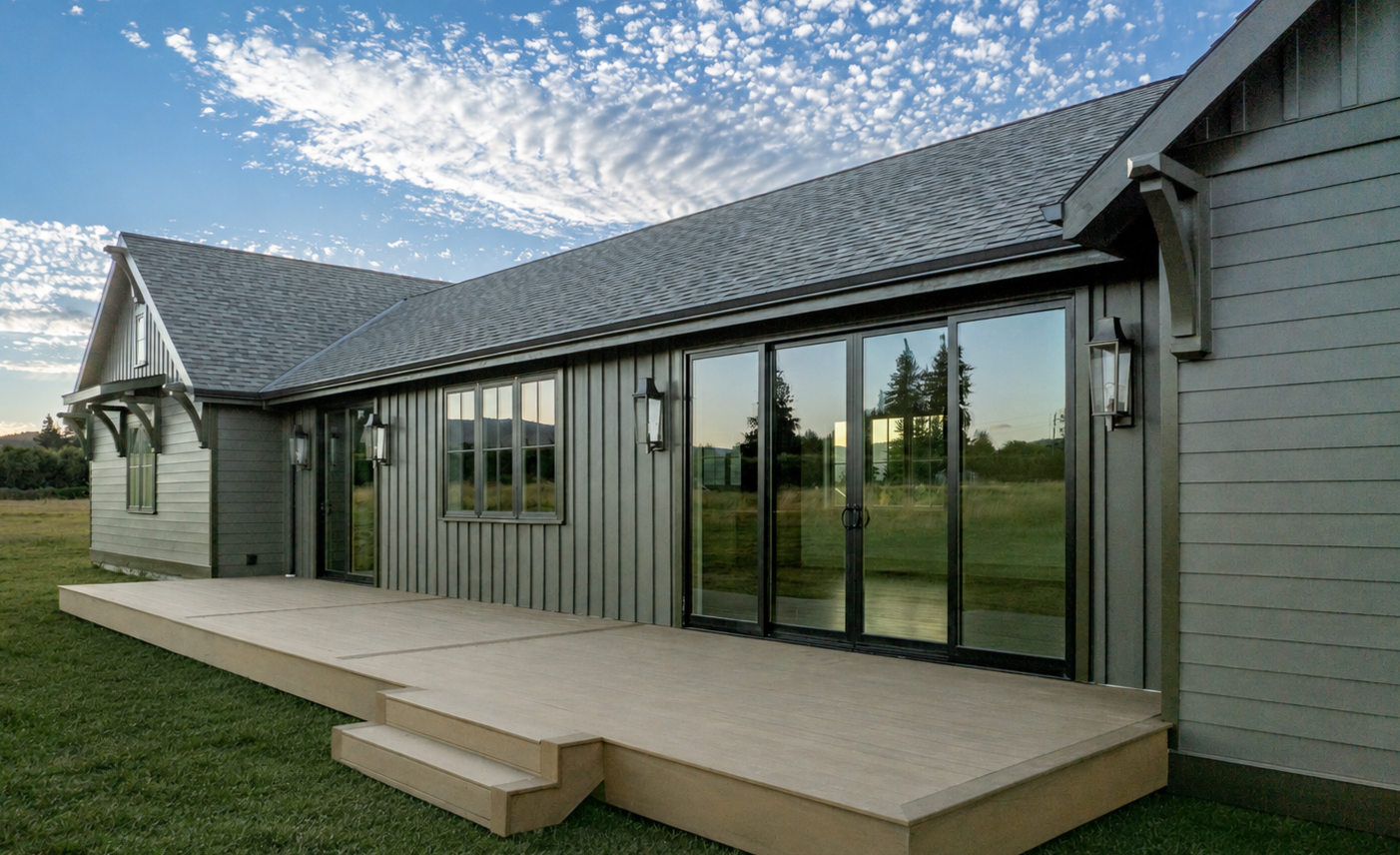 Transitional traditional style new build house with gray slate colored siding, large dark bronze glass sliding doors, a wooden deck, and outdoor wall-mounted lantern lights, under a partly cloudy sky in Hood River.