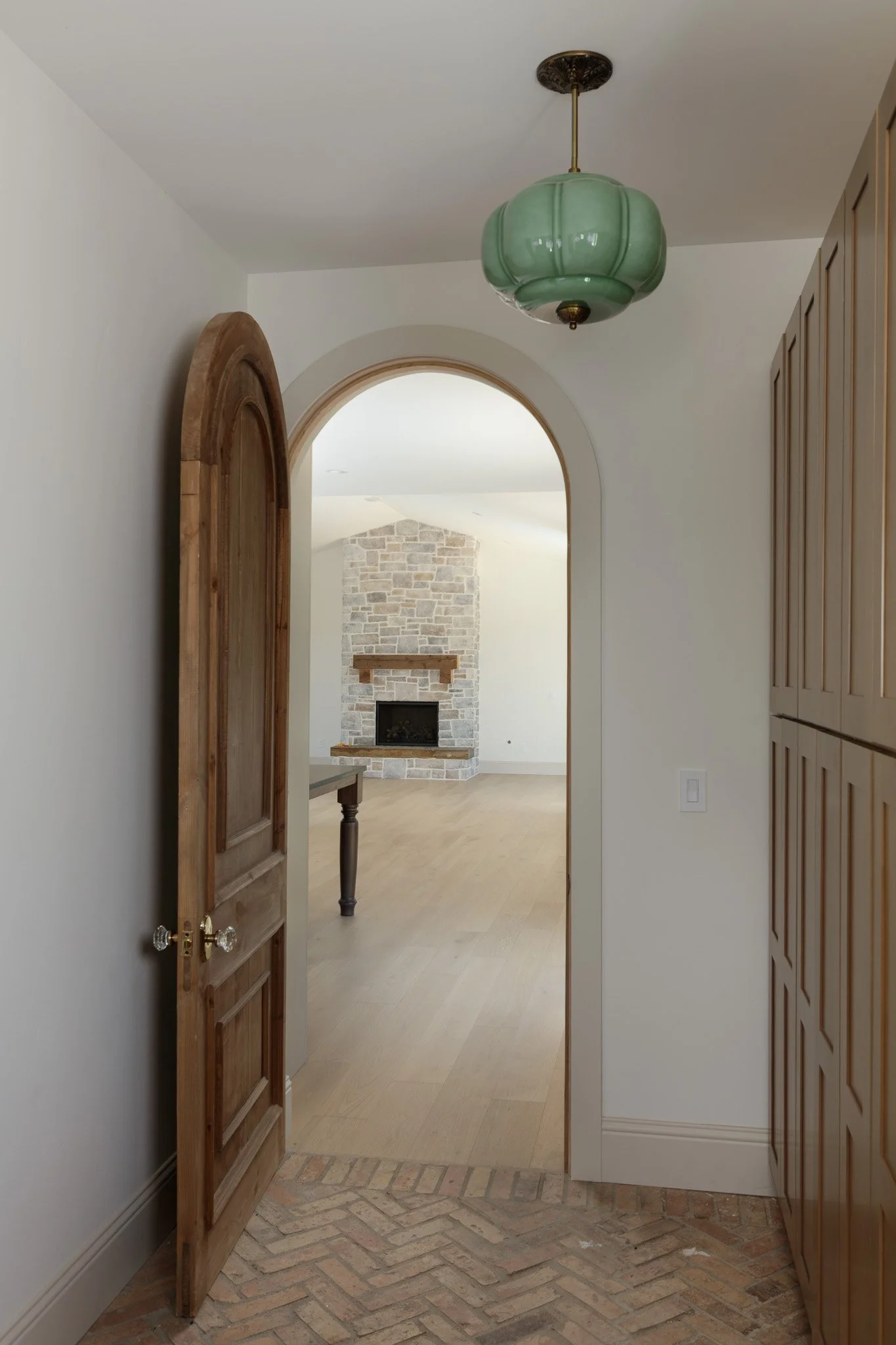 An interior hallway with a wooden door open to a room featuring a stone fireplace, light wood flooring, built-in wooden cabinet, and a green vintage hanging lantern ceiling light.