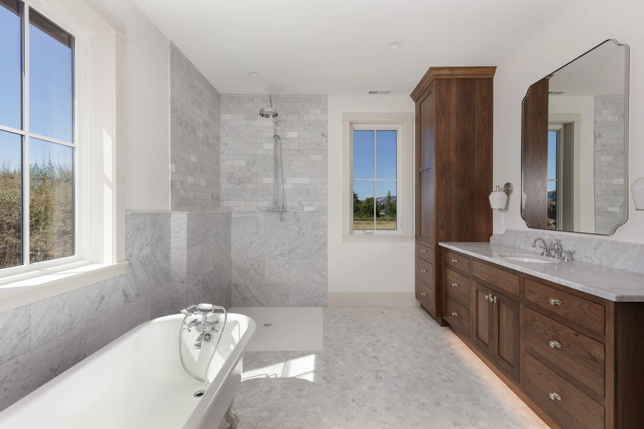 New build custom bathroom with large windows, bathtub, white marble walk-in shower with marble tile walls, custom inset cabinetry wooden vanity with marble countertop, and large mirror.