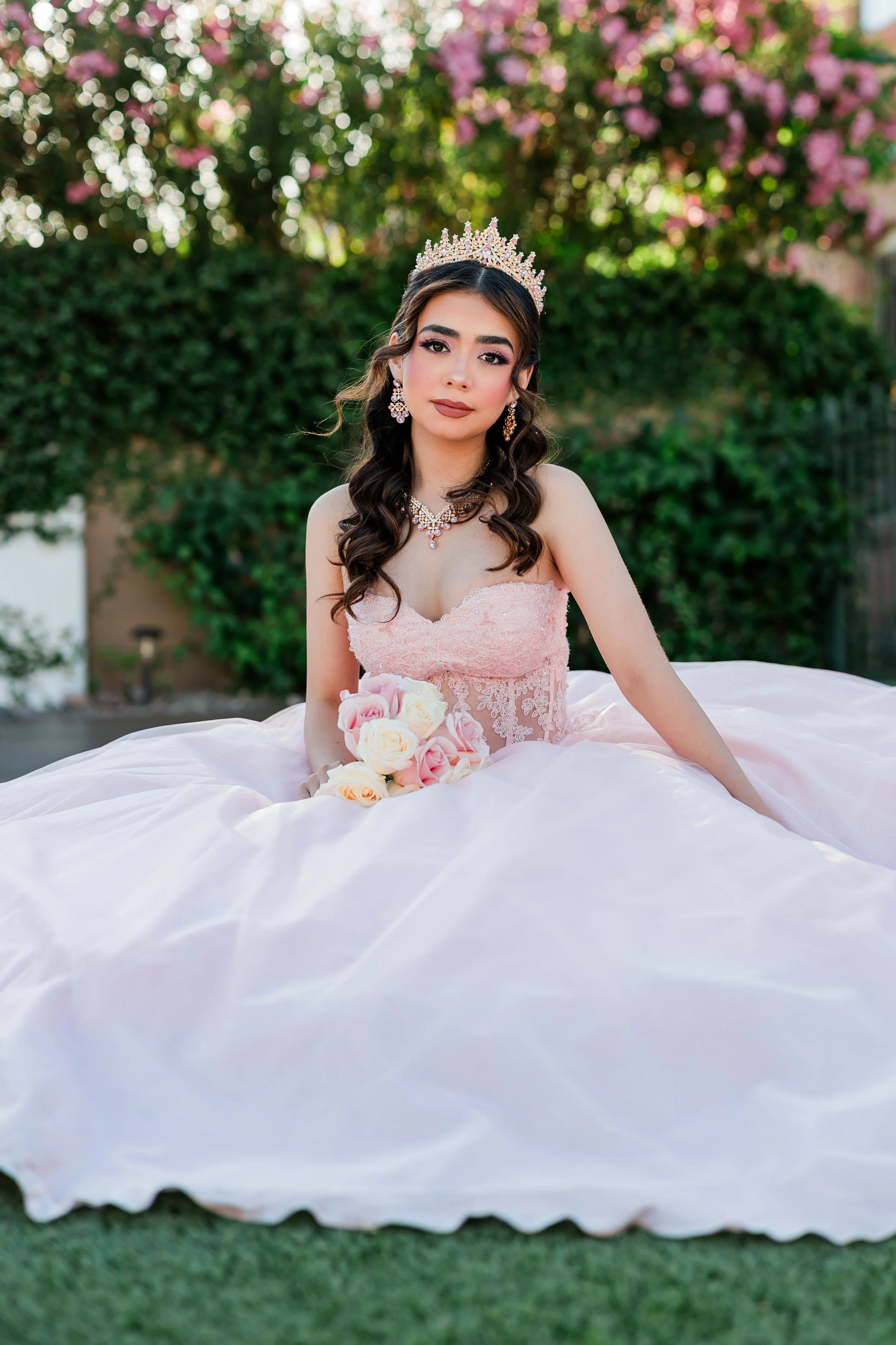 Young woman in a pink lace strapless dress sitting on a grassy lawn with a bouquet of pink and white roses, wearing a tiara, jewelry, and styled hair, outdoors with a bush and pink flowering tree in the background.
