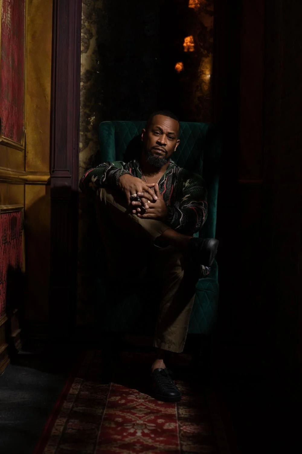 Tahoe with in dark green chair, dark background, antique room in dark blazer and bottoms, legs crossed