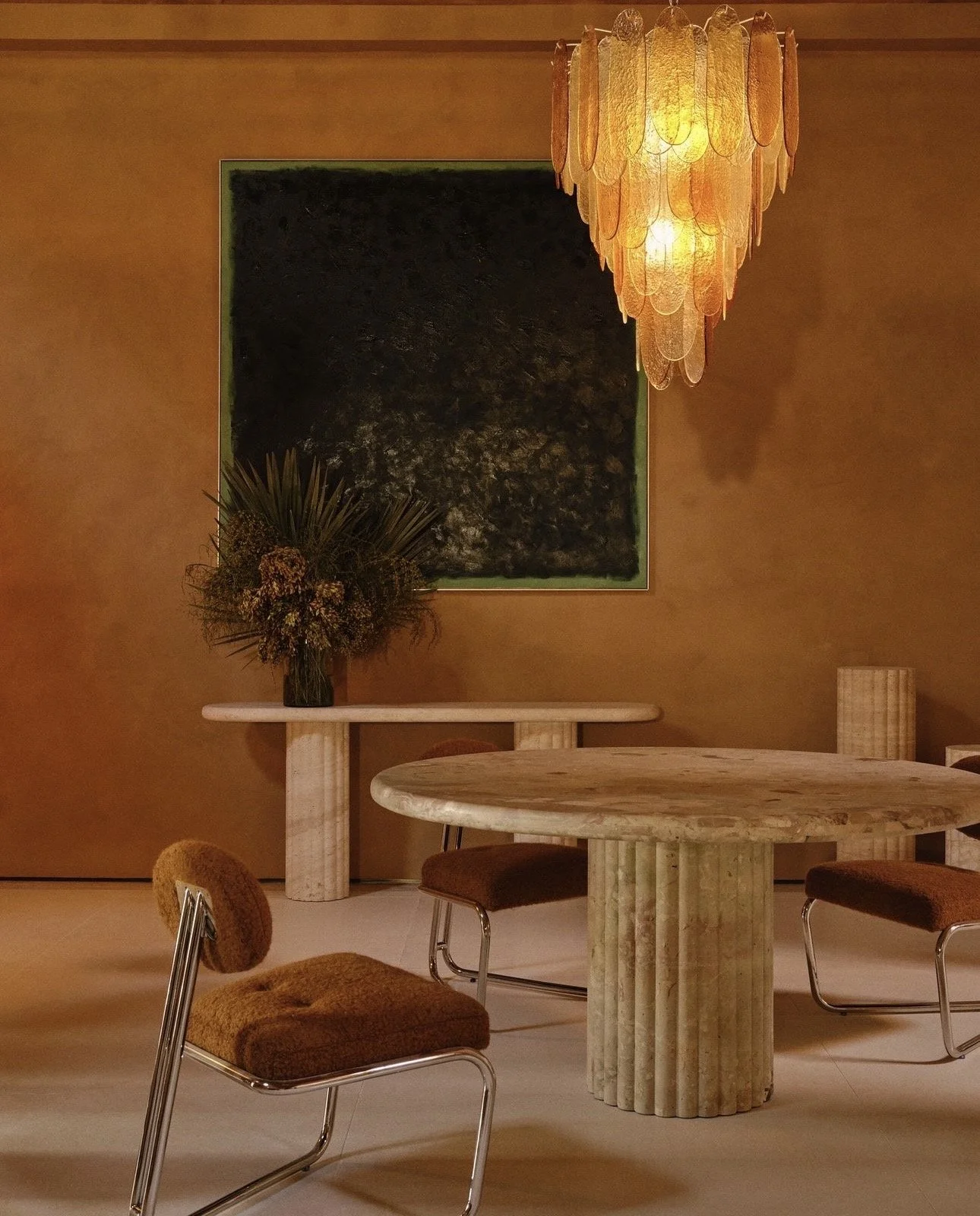 Interior of a room with a round marble table, three brown chairs with chrome legs, a rectangular marble console table with a floral arrangement, an abstract black painting on the wall, and an orange pendant chandelier.