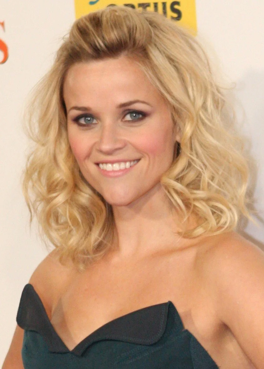 Headshot of Reese Witherspoon with short blonde curls and an angular dress neckline.