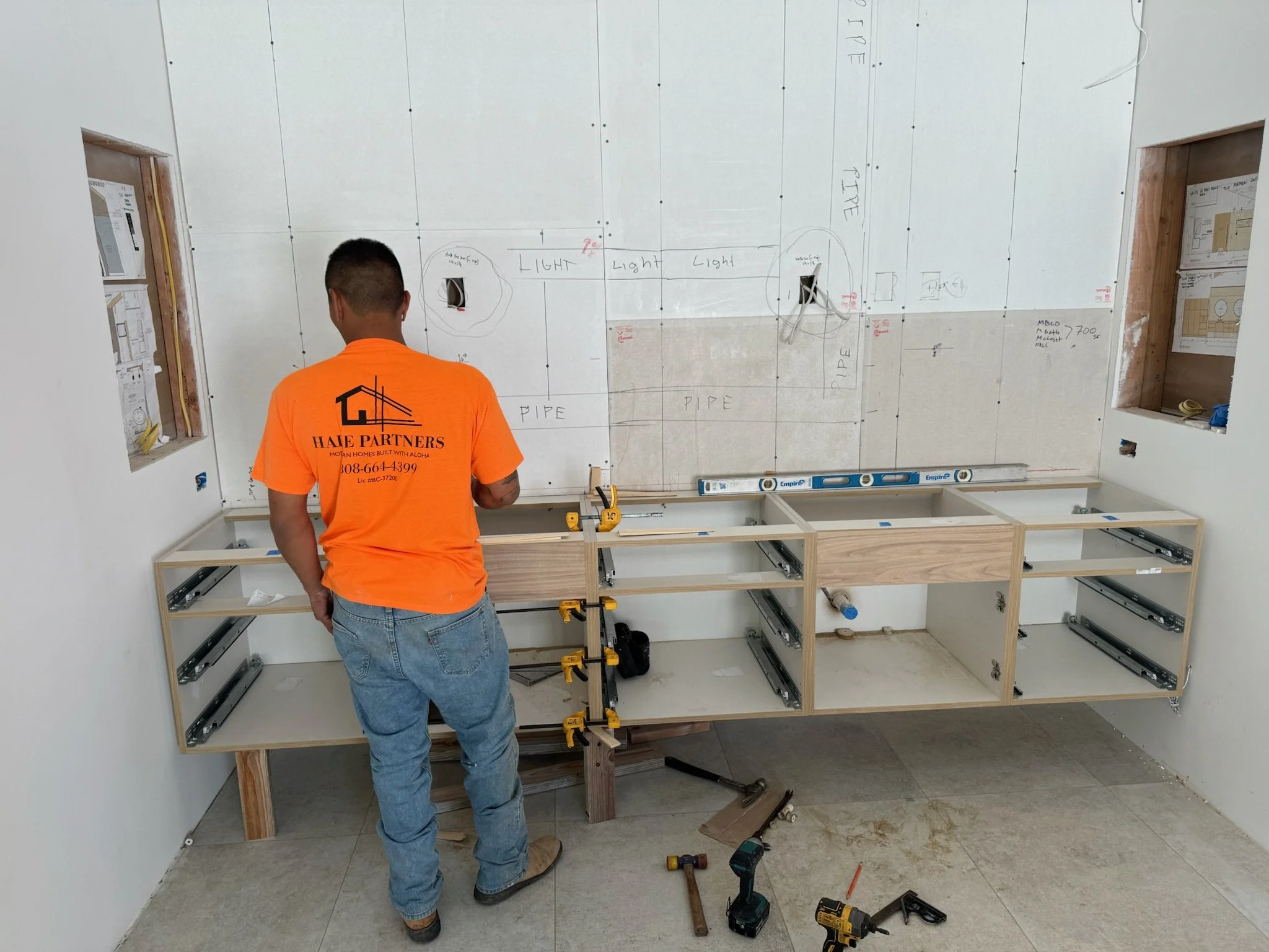 Construction worker installing a wooden cabinet frame on a wall in a room. The wall has markings and sketches for plumbing and electrical installations.
