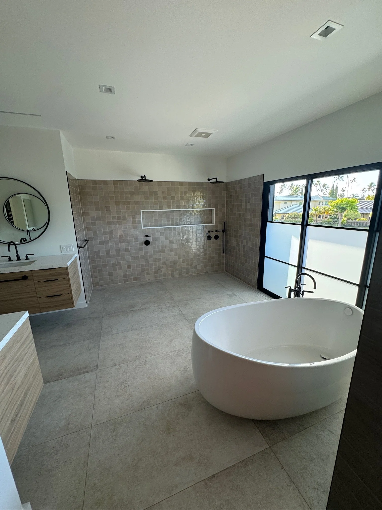 Modern bathroom with large freestanding bathtub, shower area with separate showerheads, vintage-style vanity with circular mirror, large window with frosted glass, neutral color palette, and minimal decor.