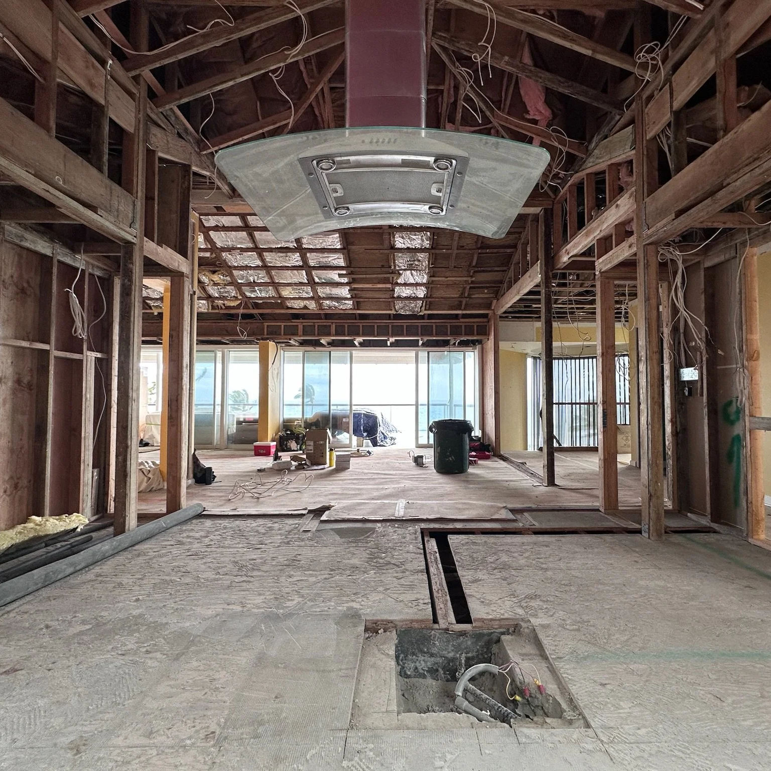 Interior of a building under renovation, showing exposed wooden framing, electrical wiring, and construction materials, with large windows in the background.