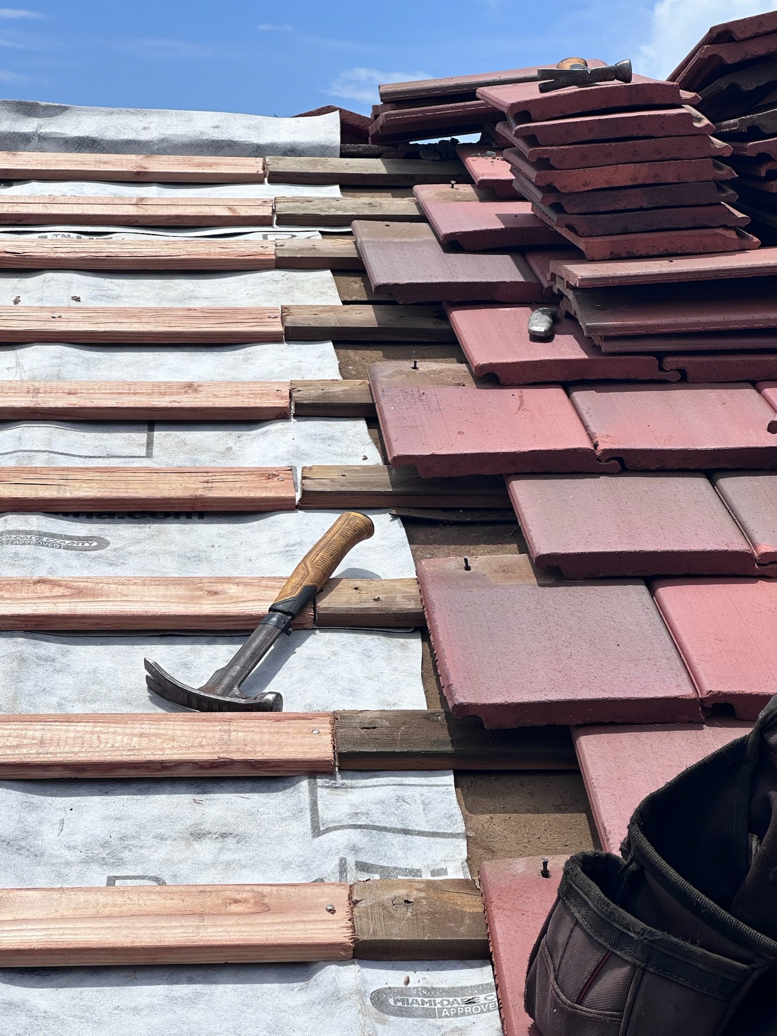 Roof construction with wooden and red tiles, a hammer on the wooden base, and a tool bag on the right side under a clear blue sky.
