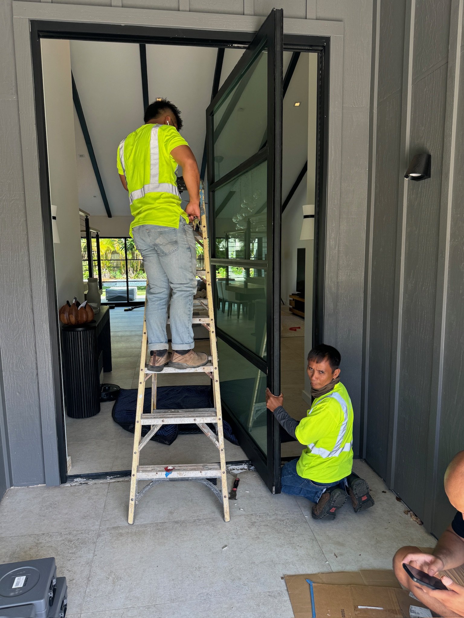 Two workers installing or repairing a large glass door or window at the entrance of a modern house, one standing on a ladder and the other kneeling on the ground.