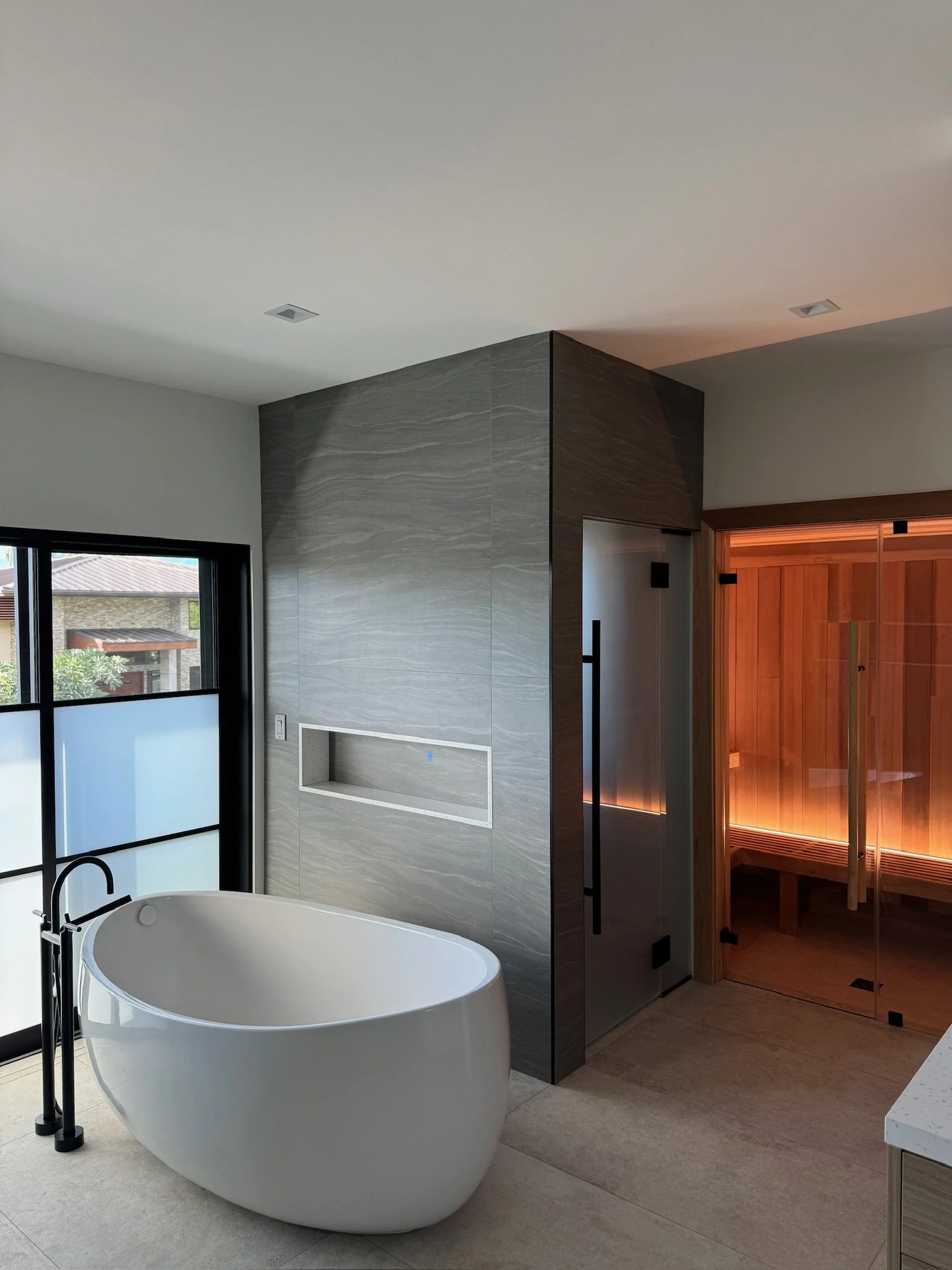 Modern bathroom featuring a white freestanding bathtub with a black floor-mounted faucet, large window with frosted and clear glass panels, gray tiled wall, and a wooden sauna with glass door inside.
