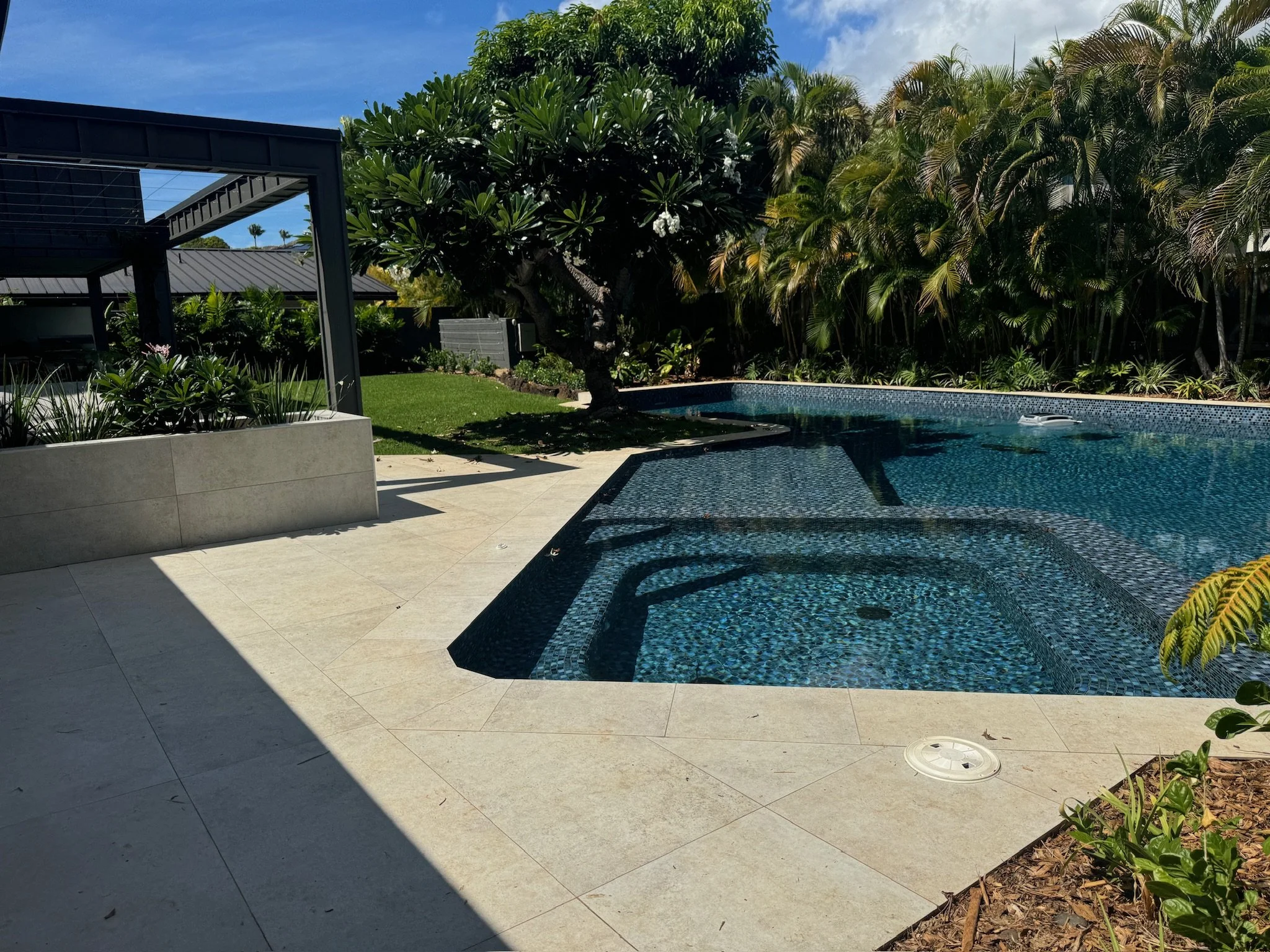 A modern backyard with a swimming pool surrounded by tropical plants and trees. There is a shaded patio area with a metal framework on the left, and the pool features built-in seating and steps. Bright sunlight casts shadows on the beige tiled pool d