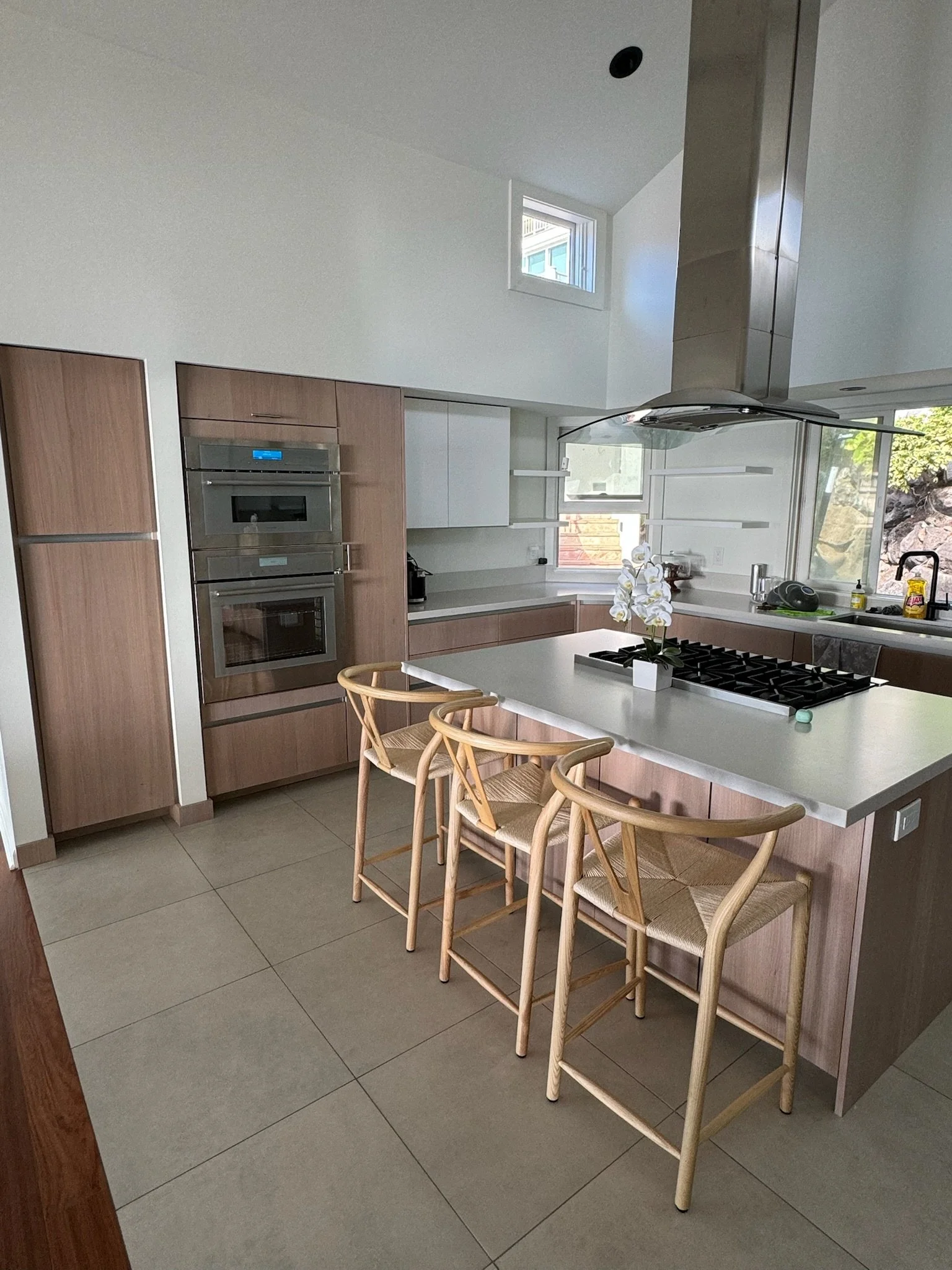 Modern kitchen with white countertops, wooden cabinets, a built-in oven, a stainless steel range hood, a small window, and three wooden bar stools at the island.