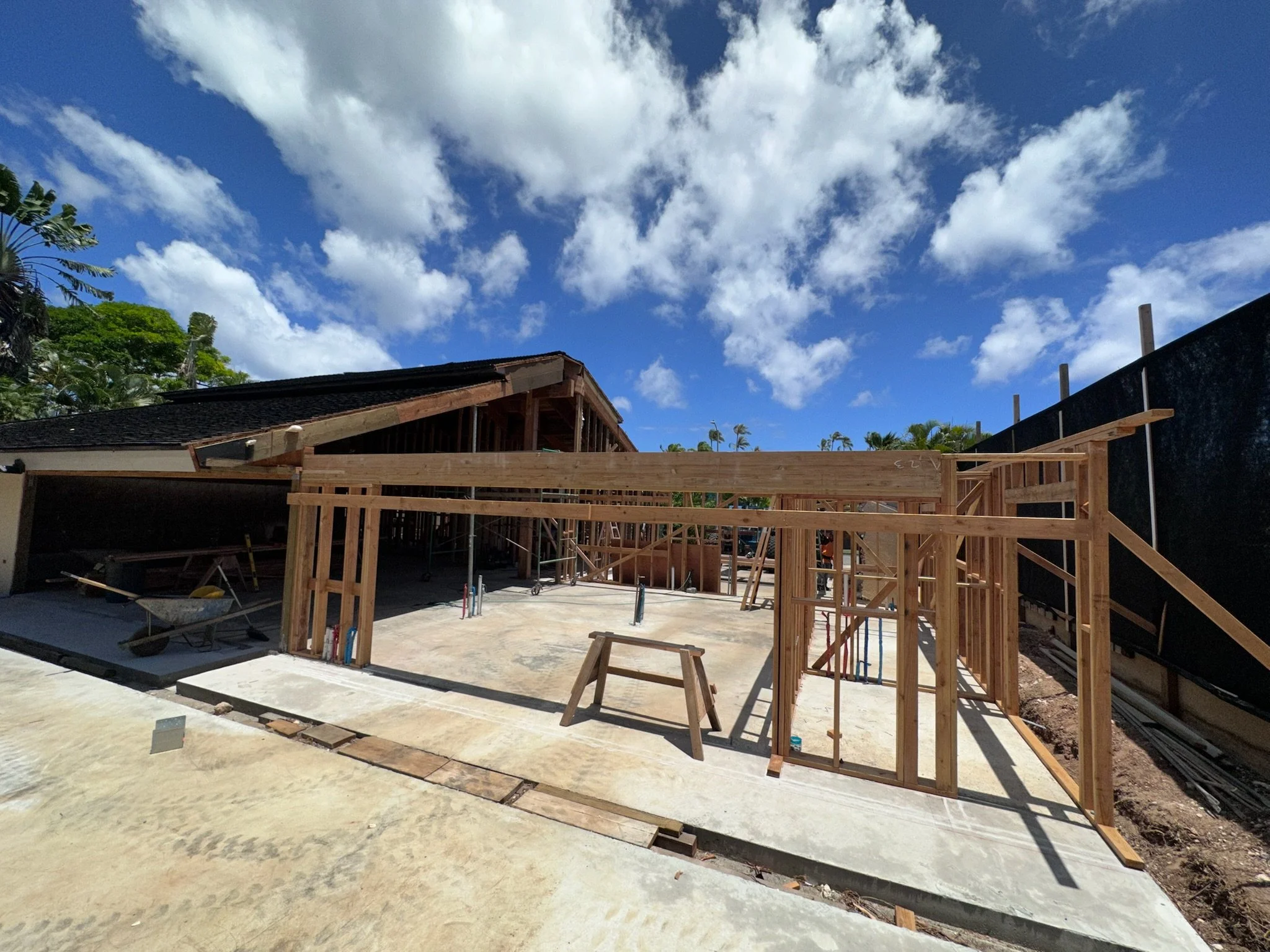Construction site with wooden framing under a blue sky with white clouds, concrete foundation, and scaffolding in the background.
