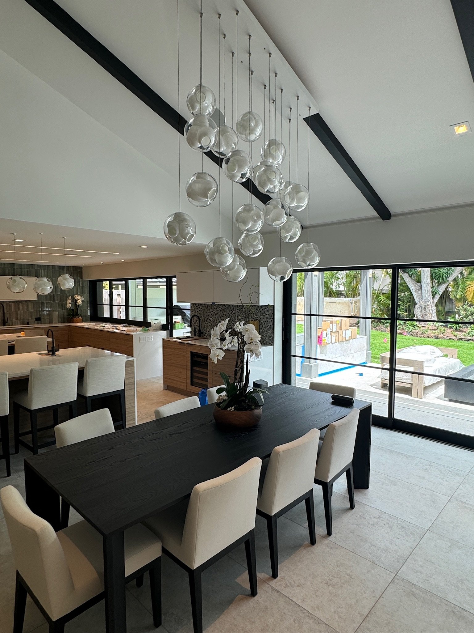 Modern dining room with a black table, cream chairs, a floral centerpiece, large glass pendant chandelier, and sliding glass doors leading to a backyard.