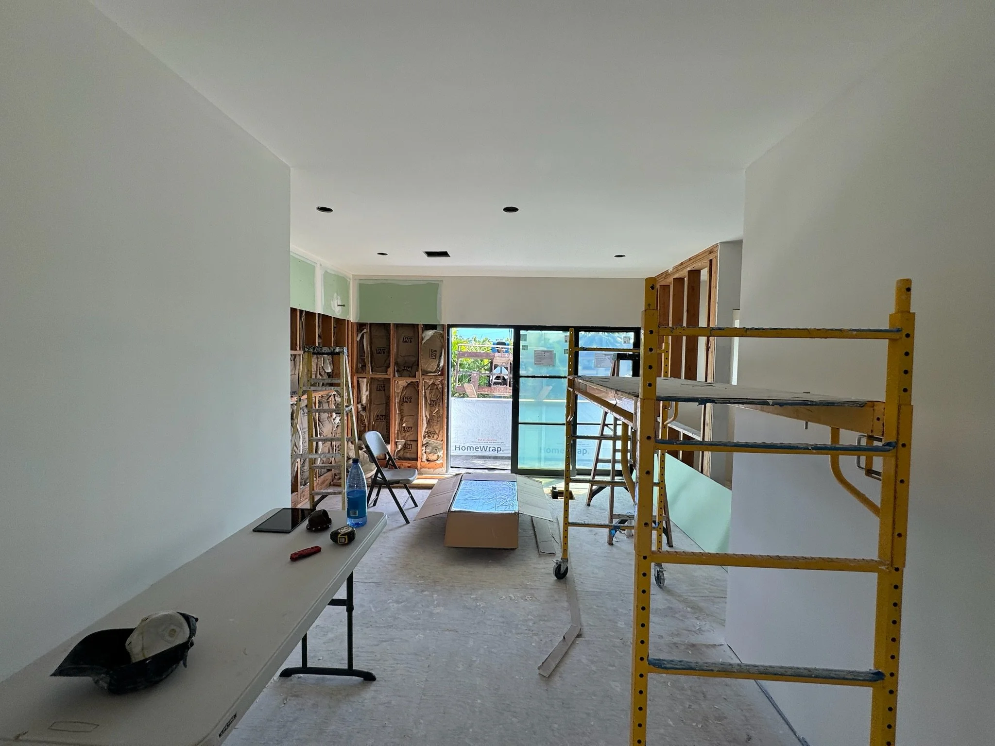 Interior of a room under construction with drywall installation in progress, a yellow scaffold, a folding chair, tools on a table, and a sliding glass door leading outside.