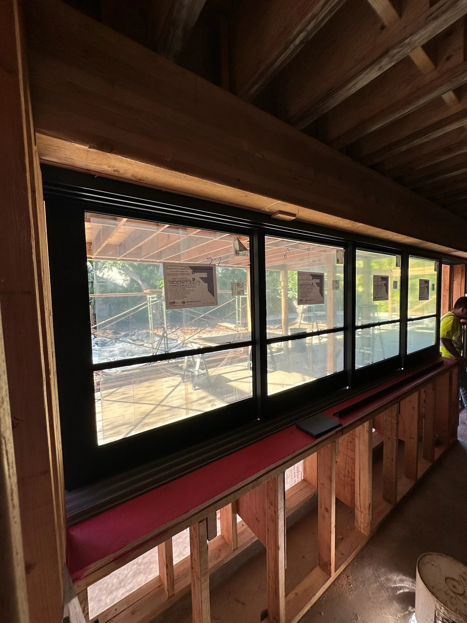 Interior of a building under construction with a large black-framed window showing a construction site outside. Wooden framing and a worker wearing a yellow vest are visible inside.