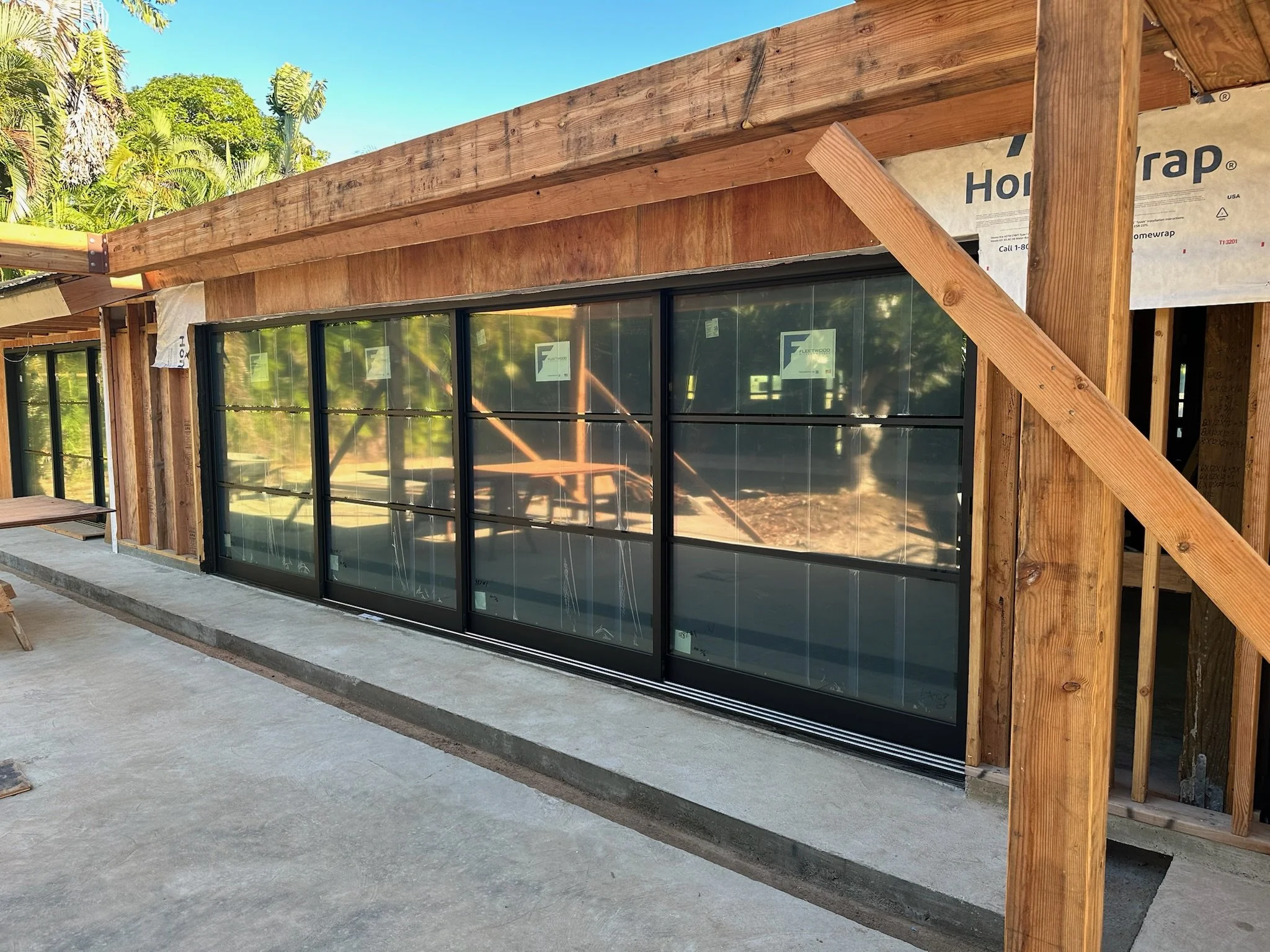Partially constructed building with large sliding glass doors, wooden framing, and concrete foundation, with tropical trees and blue sky in background.
