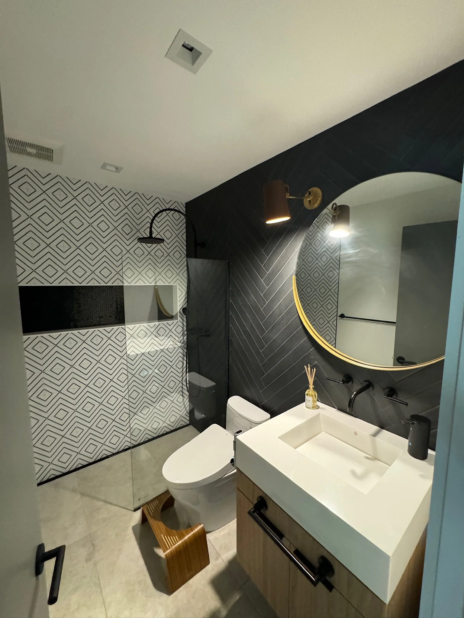 Modern bathroom with black and white geometric tile accents, a glass-enclosed shower, a white toilet, and a wooden vanity with a white sink. Decor includes a round mirror with a gold frame, a diffuser, and wall-mounted lights.