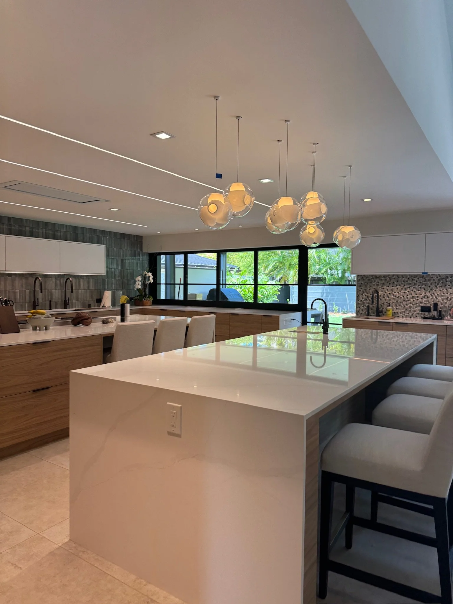 Modern kitchen with island, pendant lights, bar stools, and large window with greenery outside.