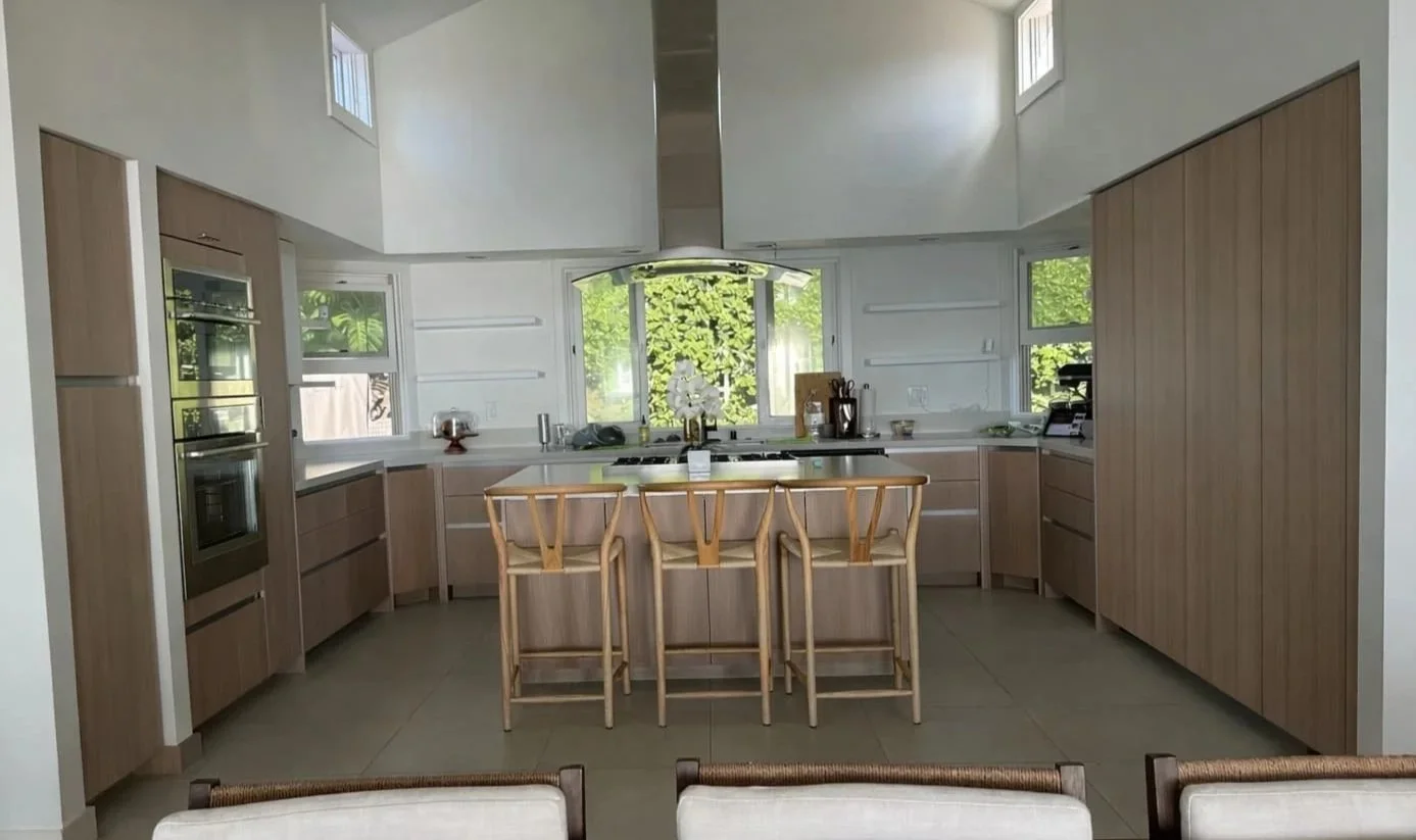 Hale Partners Hawaii's modern kitchen renovation post construction, with wooden cabinets, a center island with four bar stools, and large windows showing greenery outside.