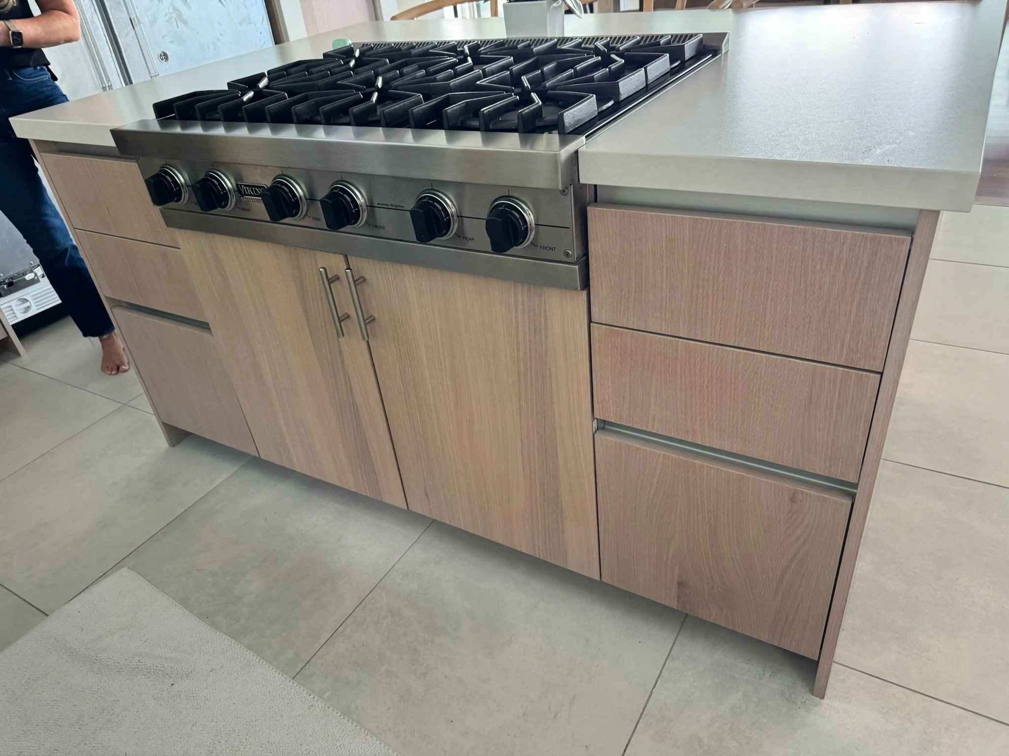A kitchen island with a built-in stovetop and oven, featuring five black gas burners and control knobs on the front, surrounded by beige cabinets and a light-colored countertop.