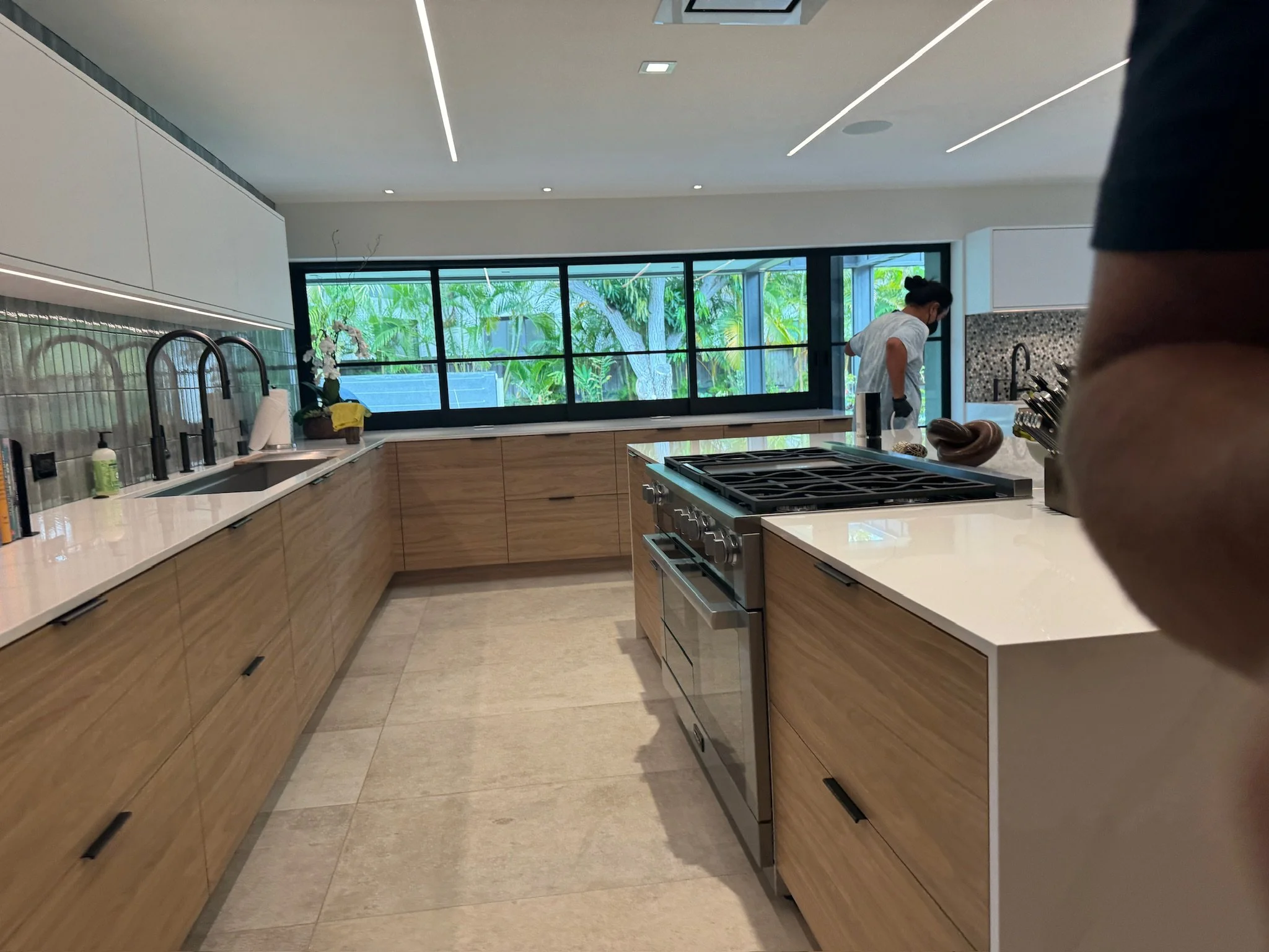 Modern kitchen with wood cabinets, a white countertop, black faucets, a large stove, and large windows overlooking greenery. A person in a white shirt is cleaning in the background.