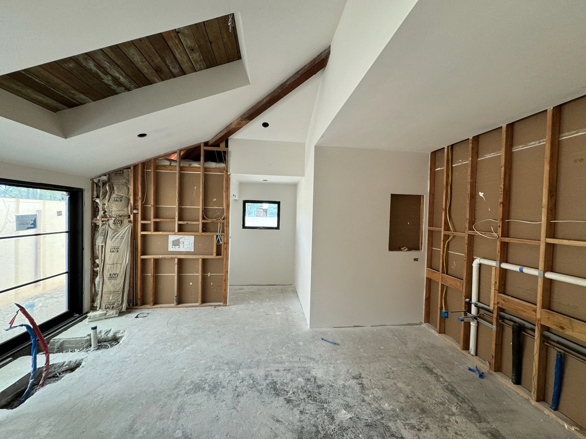Interior of a house under construction with exposed wall framing, partially installed drywall, and visible electrical wiring, with a large window on the left and a small window in the back.