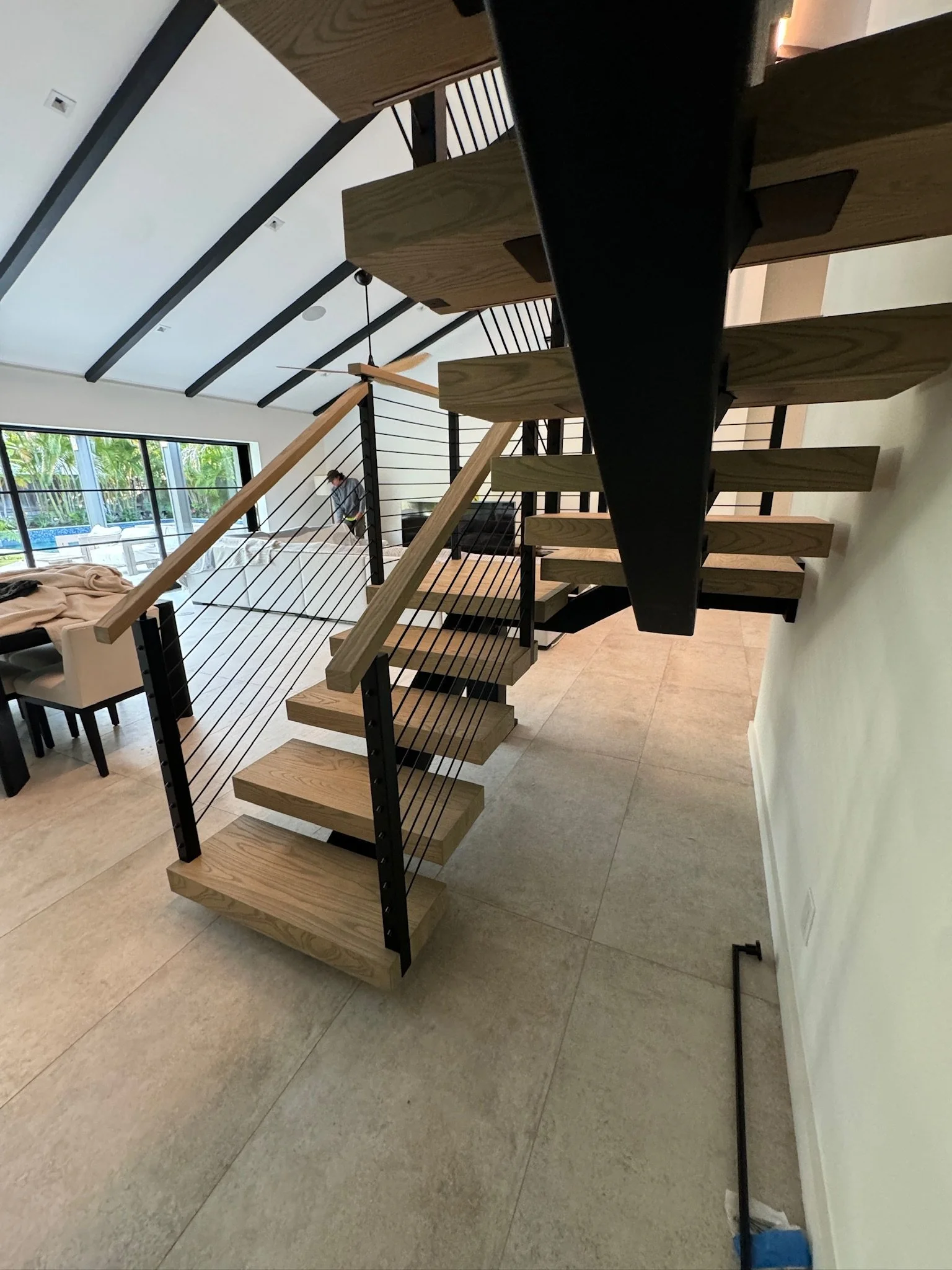 Interior view of a modern open-concept living space featuring a floating wooden staircase with black metal supports and horizontal cable railings, beige tiled flooring, and large windows showing outdoor greenery.