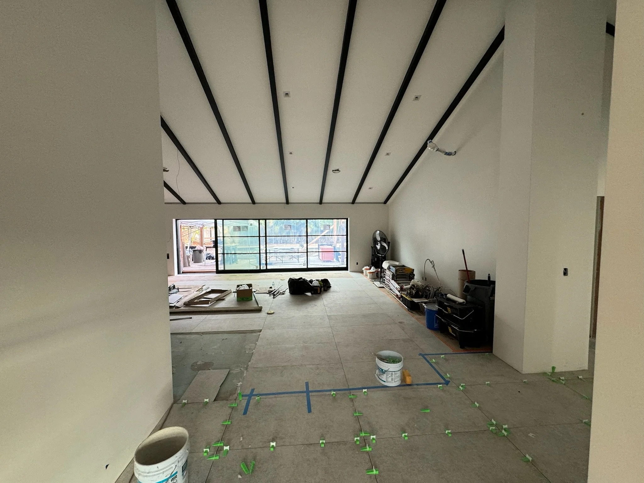 Interior of a room under construction, with visible ceiling beams, large sliding glass doors at the far end, construction tools and materials scattered on the floor, and markings for flooring layout.