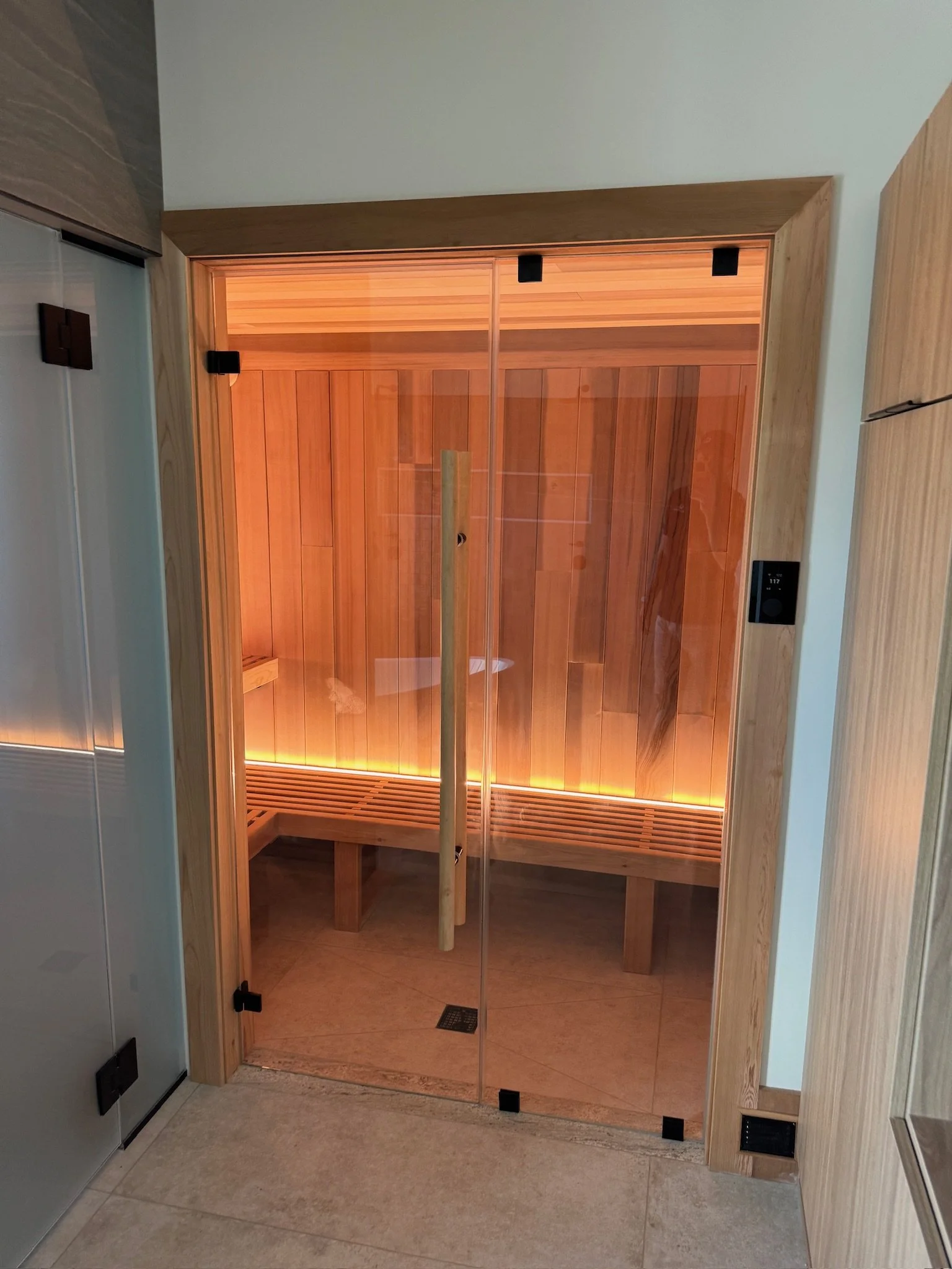 Glass door sauna interior with wooden walls, bench, and warm lighting.