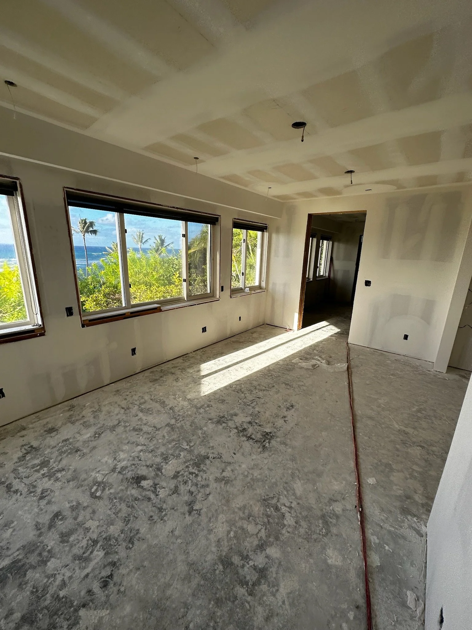 Interior of a house under construction with unfinished floors, drywall, and large windows showing a view of palm trees and the ocean.