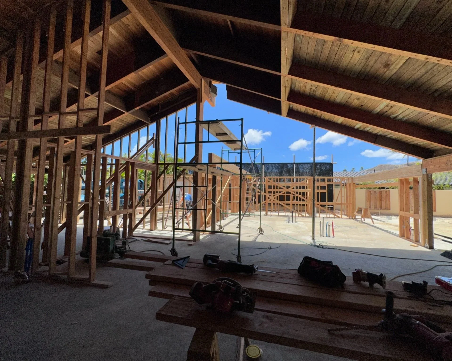 Interior view of a building under construction with exposed wooden framing, scaffolding, and construction tools on a wooden table, with a clear blue sky outside.