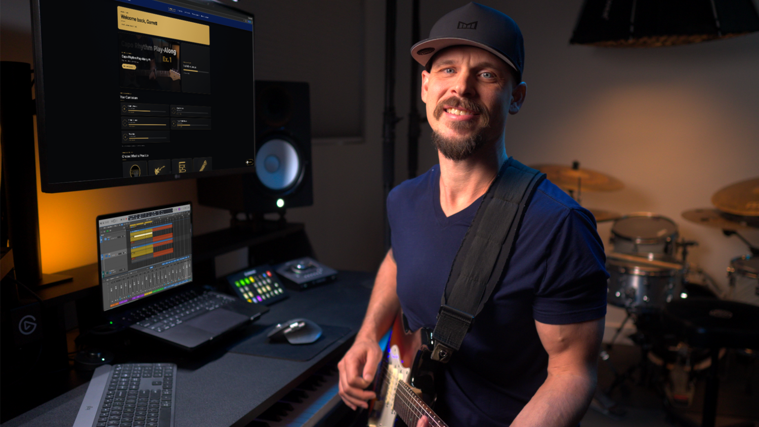 Garrett Ortego holding a guitar in his home studio, with the student dashboard and a music-production session visible on his monitors